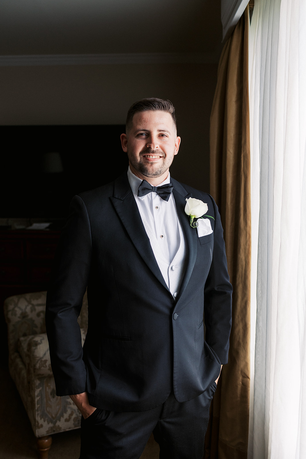 A groom in a black tuxedo stands in a window with hands in his pockets and a white boutonniere at Lambertville Station Wedding venue