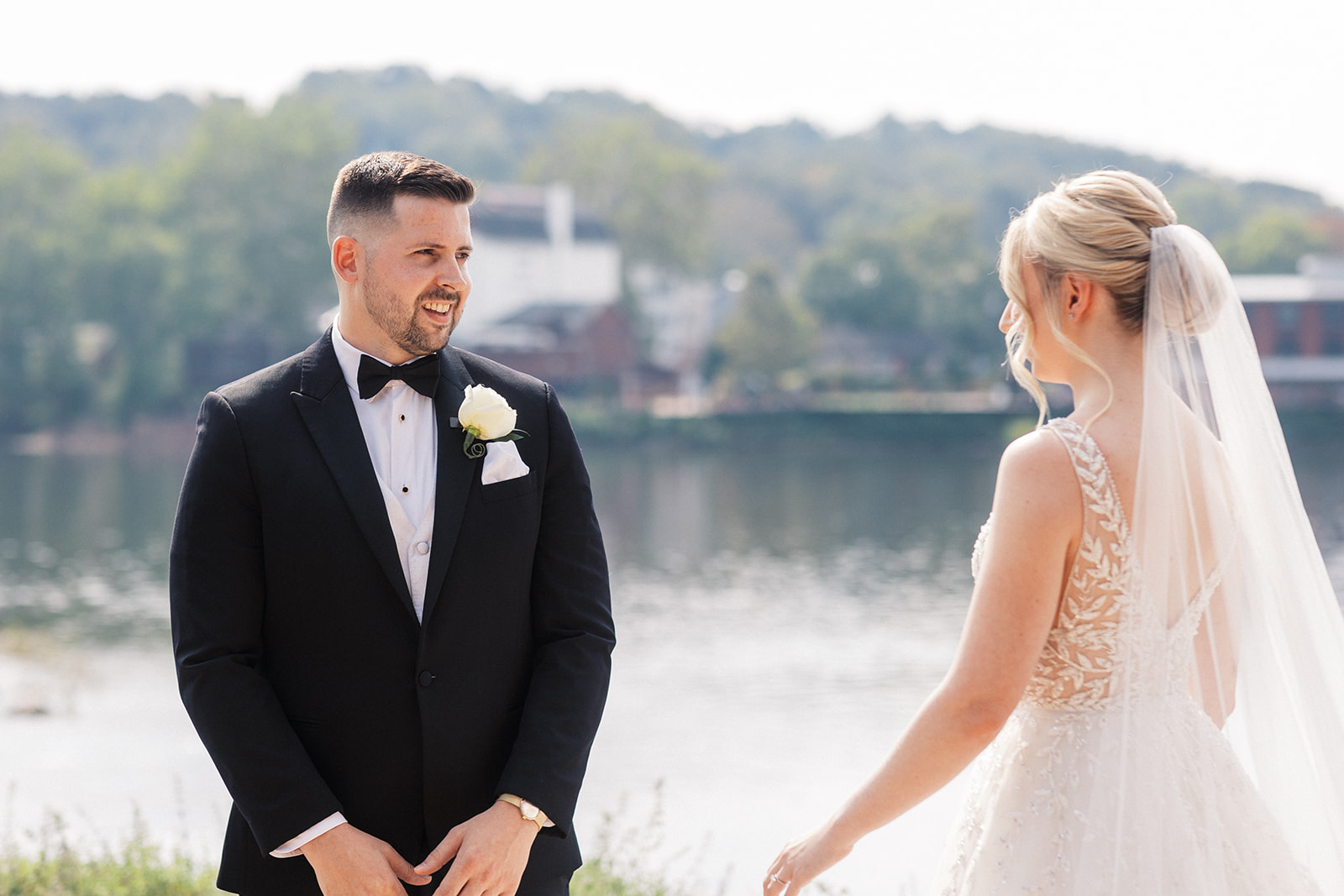 A groom in a black tuxedo smiles while standing outside by a river seeing his bride in her dress in a first look at Lambertville Station Wedding venue