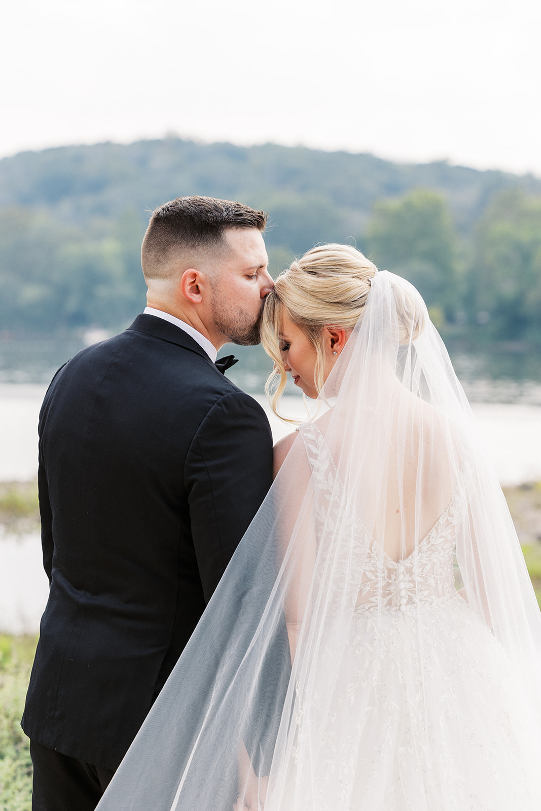 A groom kisses the forehead of his bride as they look out over the river