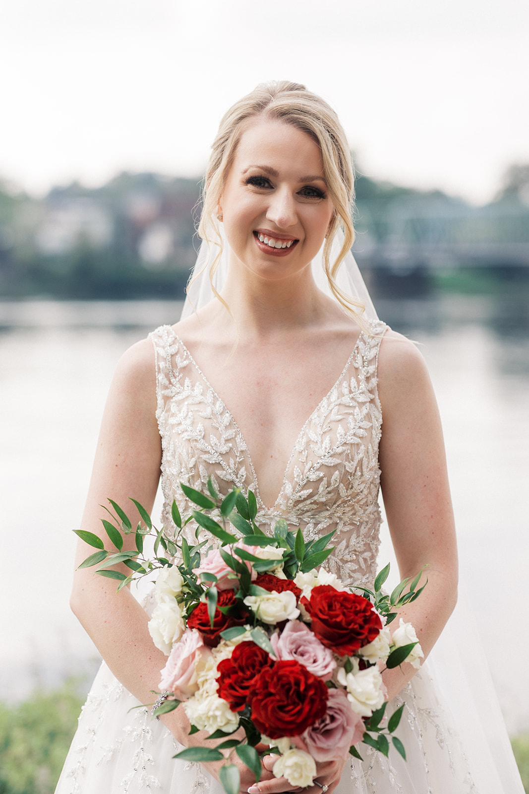 A bride stands on the riverfront at Lambertville Station Wedding venue while holding her red, pink and white bouquet