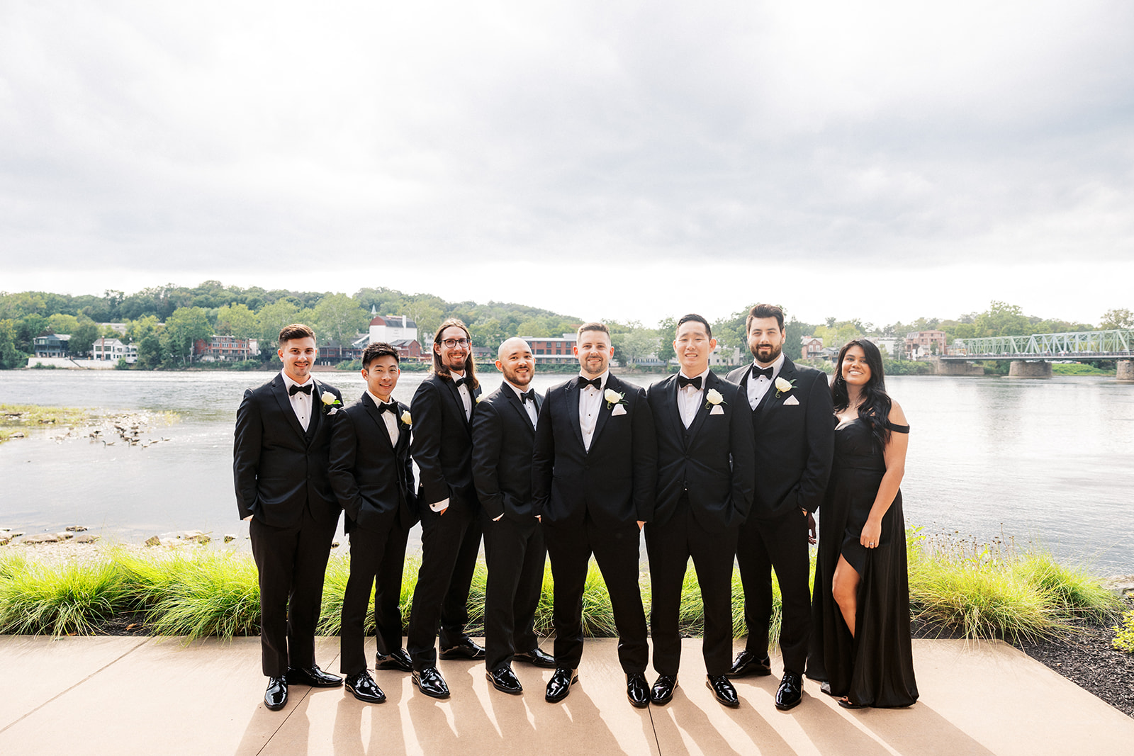 A groom in a black tux stands with hands in his pockets on a riverside patio with his large wedding party in matching suits or dress at a Lambertville Station Wedding