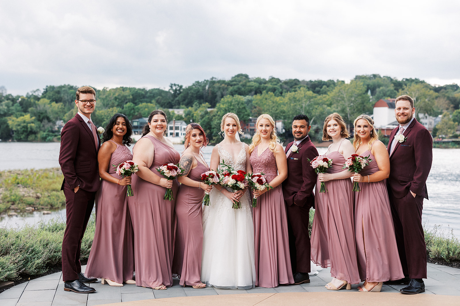A bride stands on a riverside patio holding her bouquet with her bridal party in purple suits and dresses at a Lambertville Station Wedding