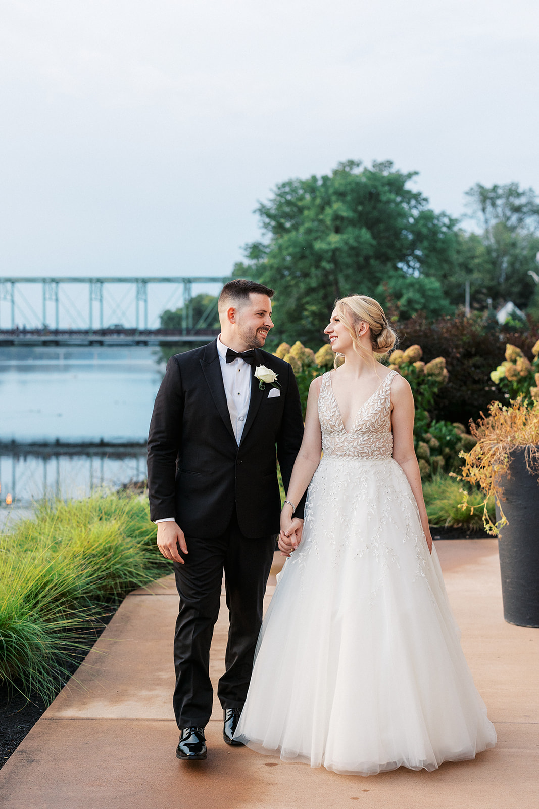 Newlyweds smile at each other while holding hands and walking through a riverfront garden