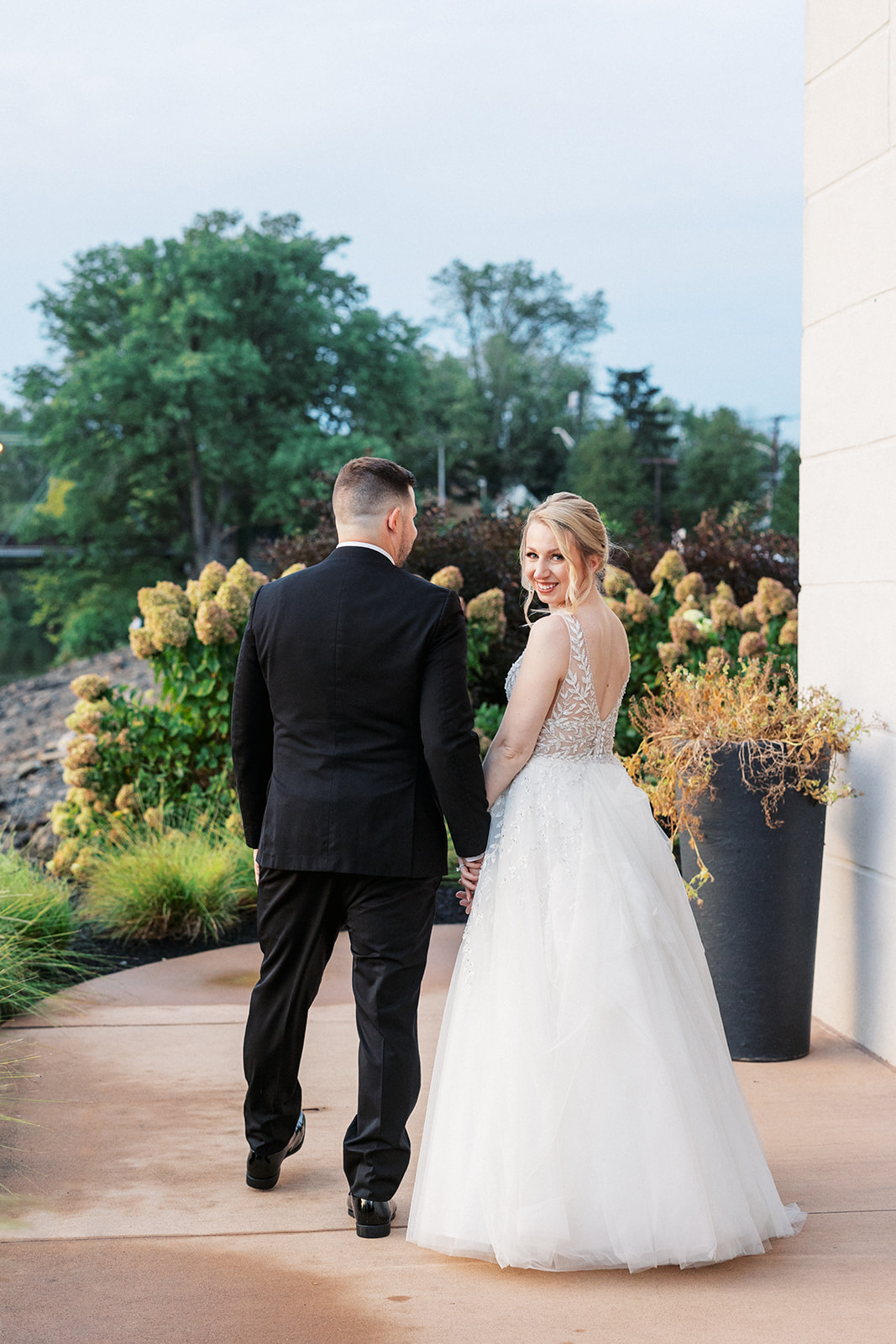 A bride looks back over her shoulder while walking hand in hand with her groom through a garden