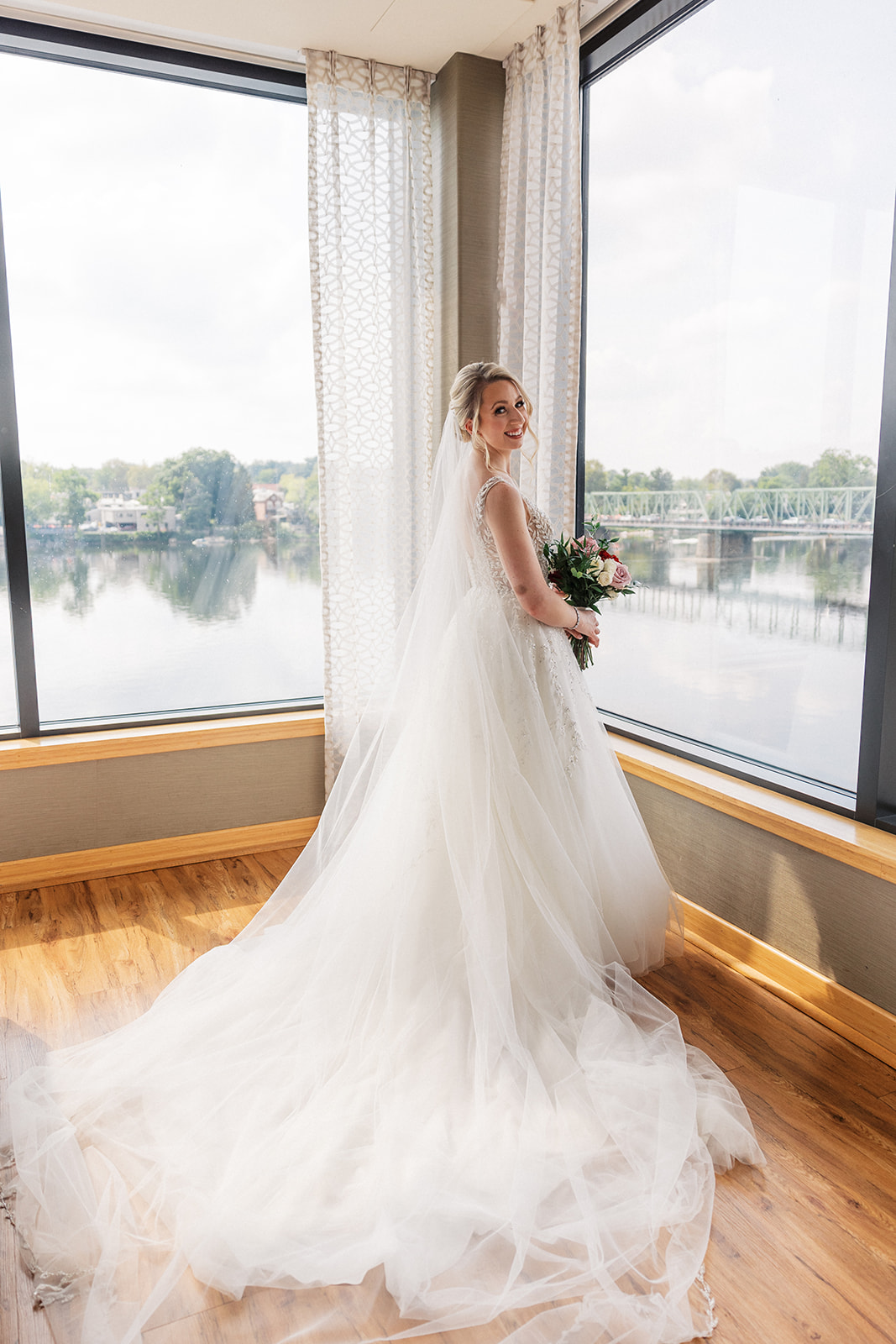 A bride stands in a corner of windows overlooking a river while holding her bouquet at Lambertville Station Wedding venue
