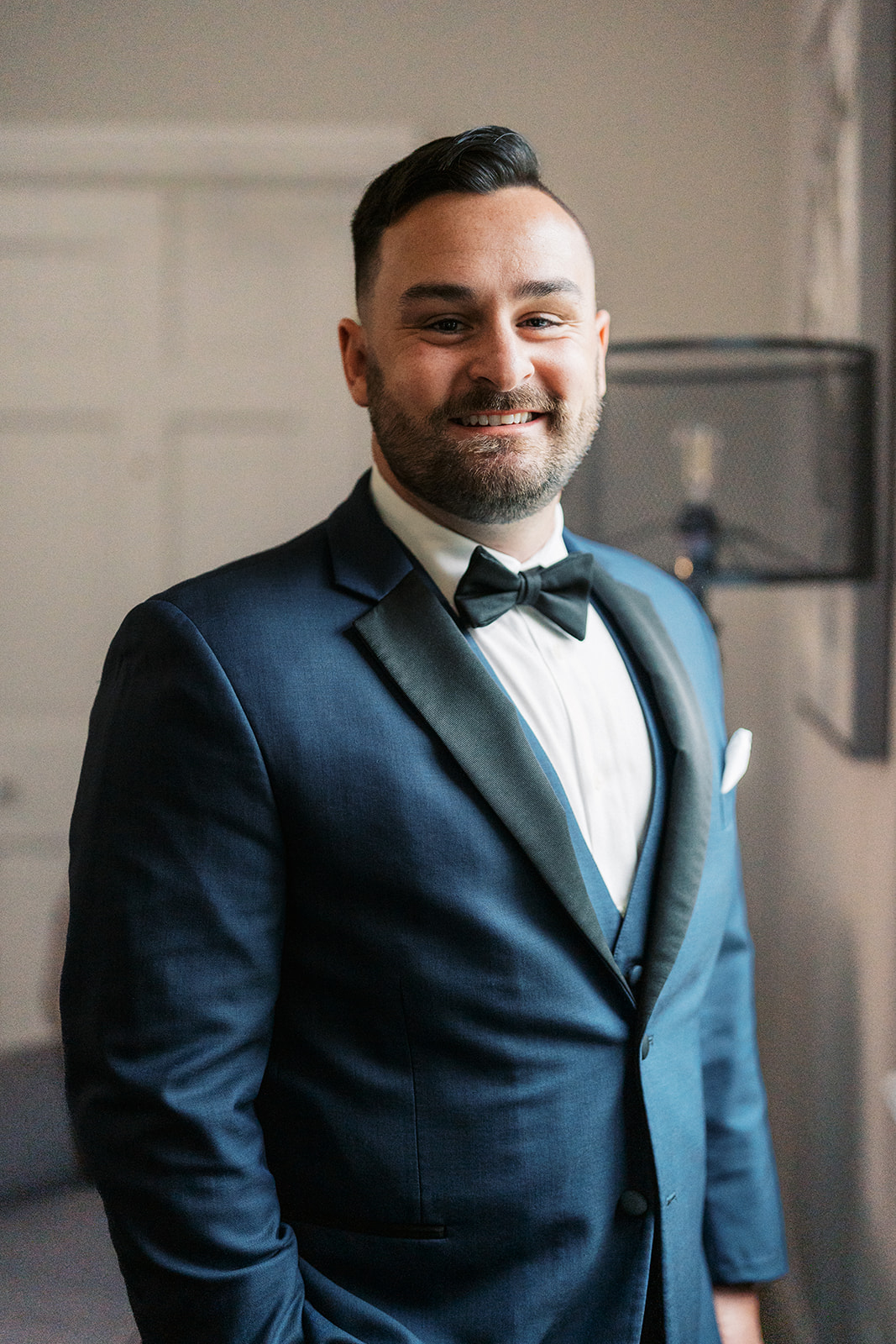 A groom smilies while standing in a window in a blue suit