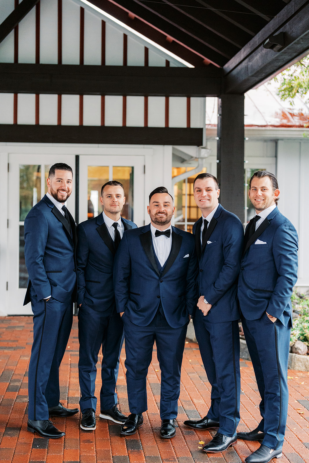A groom stands with hands in his pockets in a blue suit with his four groomsmen under a covered patio