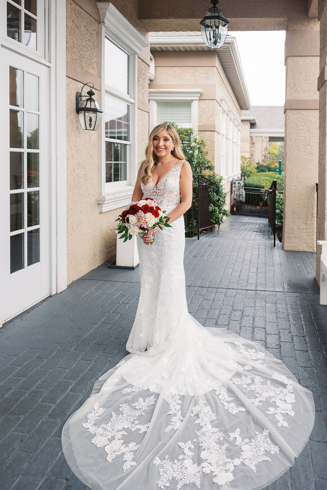 A bride in a long lace embroidered dress stands on a brick patio holding her bouquet of red and white florals