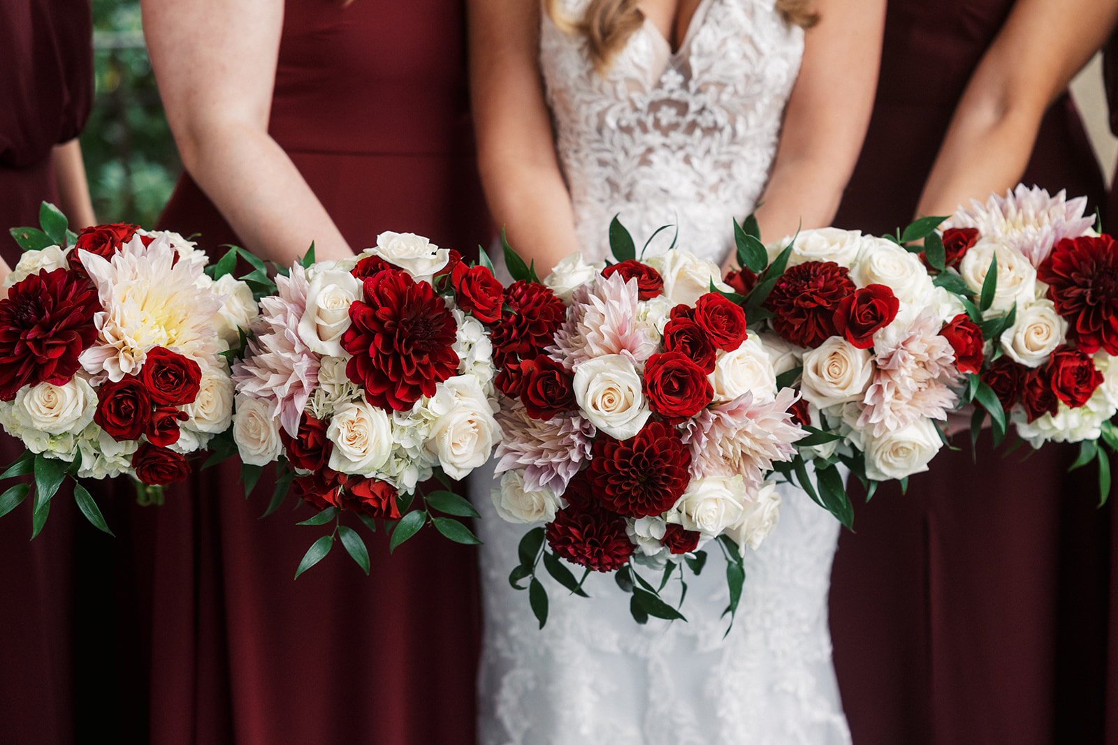 A bride stands in her lace dress holding her red and white bouquet in front of her with her bridesmaid's matching bouquets