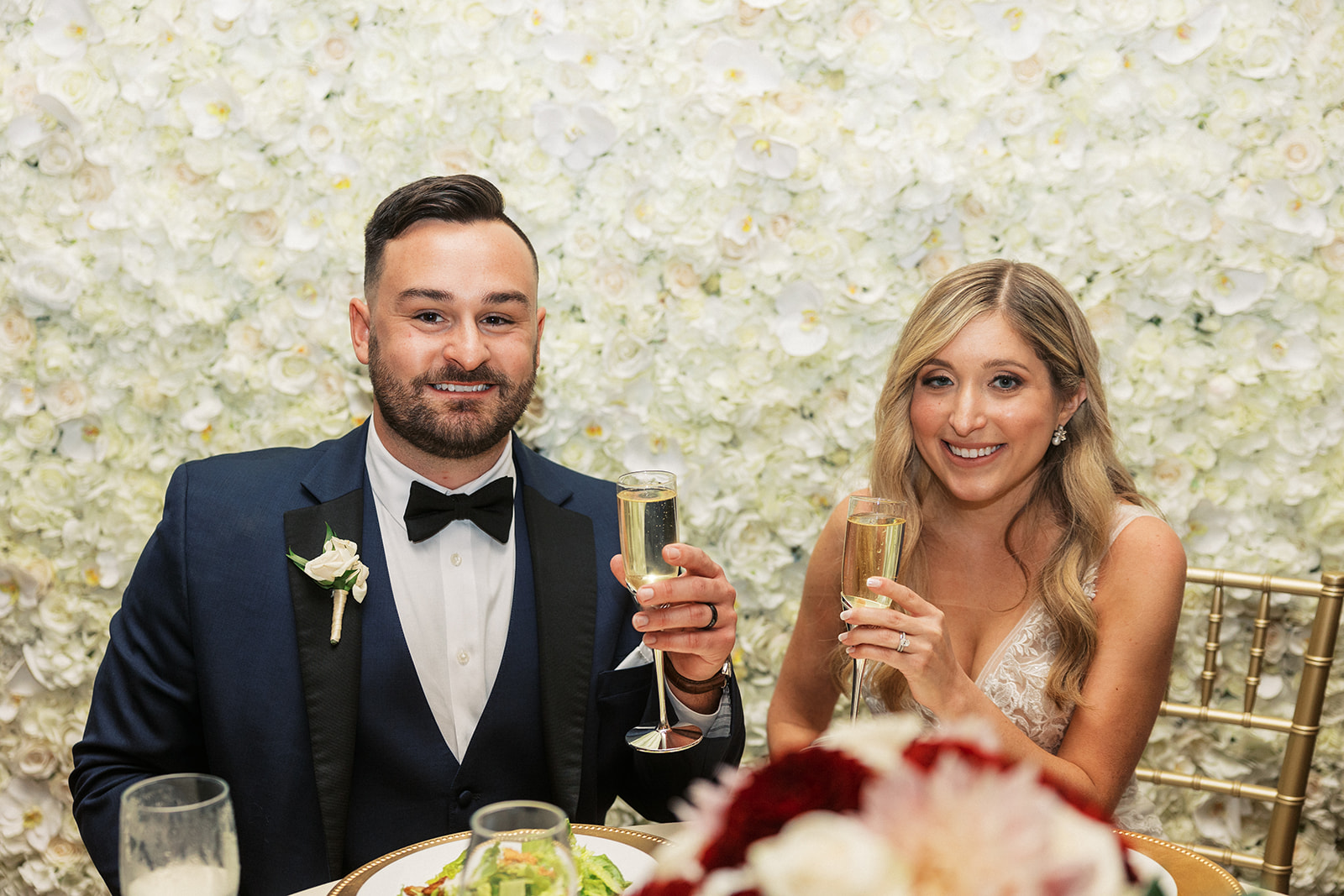 Newlyweds in a blue suit and lace dress sit at their The Belle of Blue Bell head table in front of a wall of white flowers with champagne glasses
