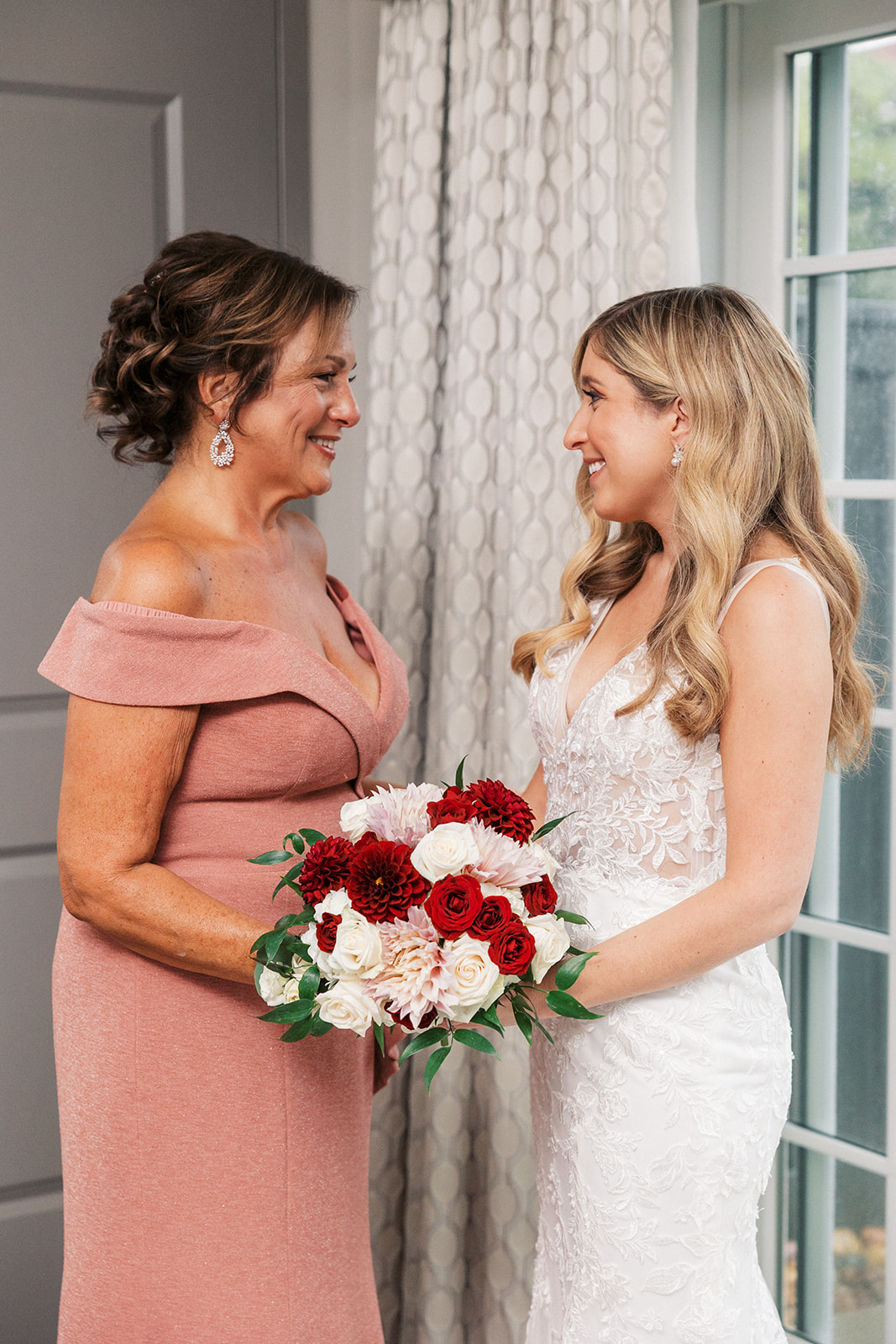 A bride smiles with her mom in the getting ready room by a window
