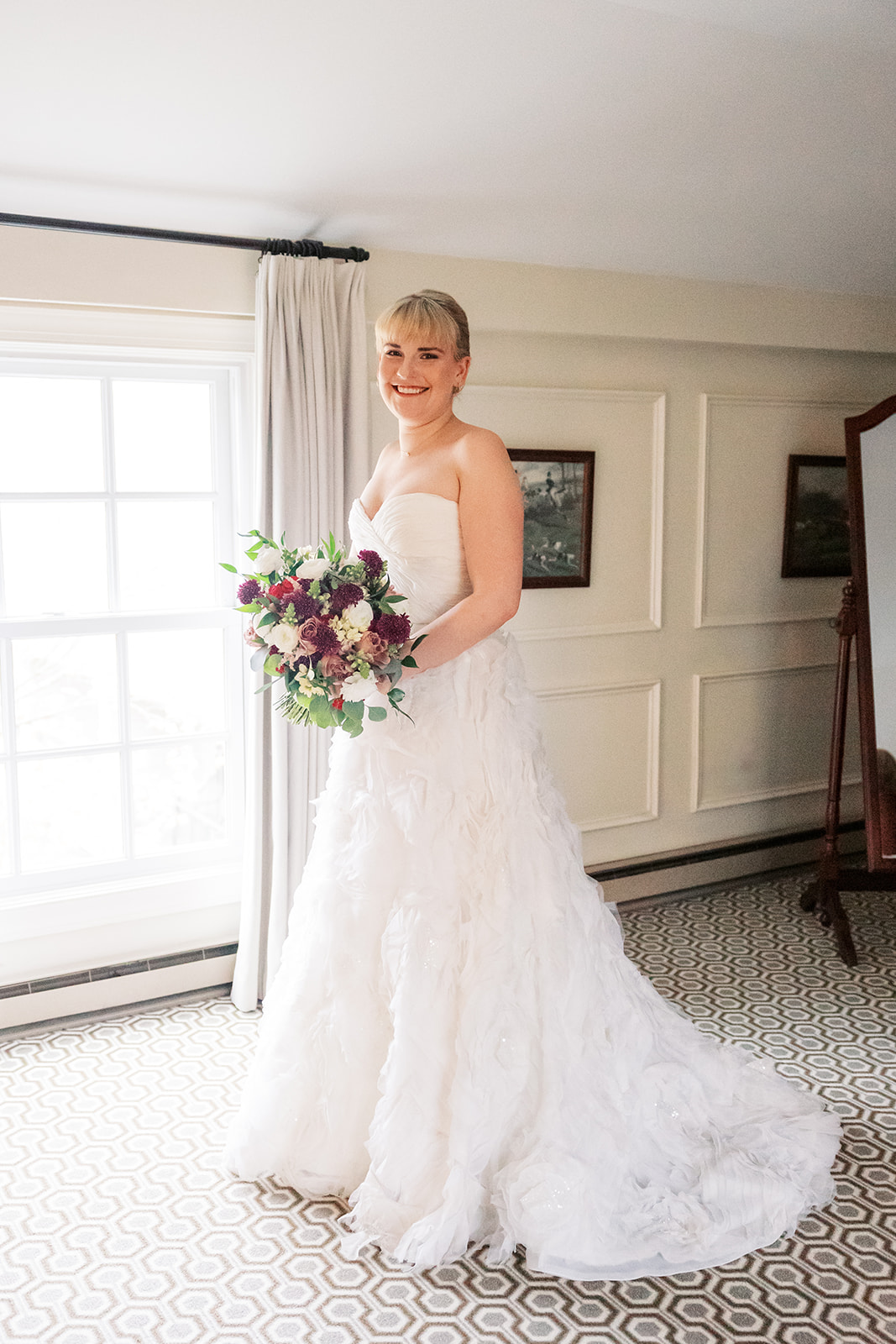 A bride stands in a window smiling and holding her bouquet