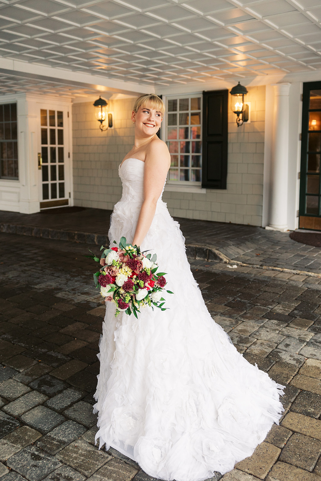A bride smiles over her shoulder while holding her colorful bouquet on a stone driveway at an Olde Mill Inn Wedding