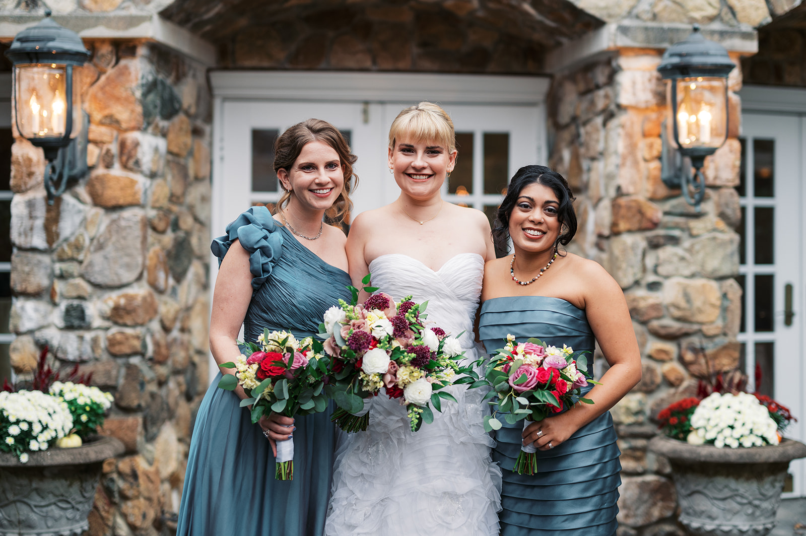 A bride stands with her 2 bridesmaids in front of stone columns holding colorful bouquets at an Olde Mill Inn Wedding