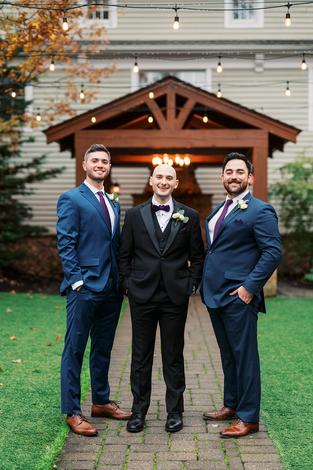 A groom stands in a black suit with 2 groomsmen in blue suits outside under market lights at an Olde Mill Inn Wedding