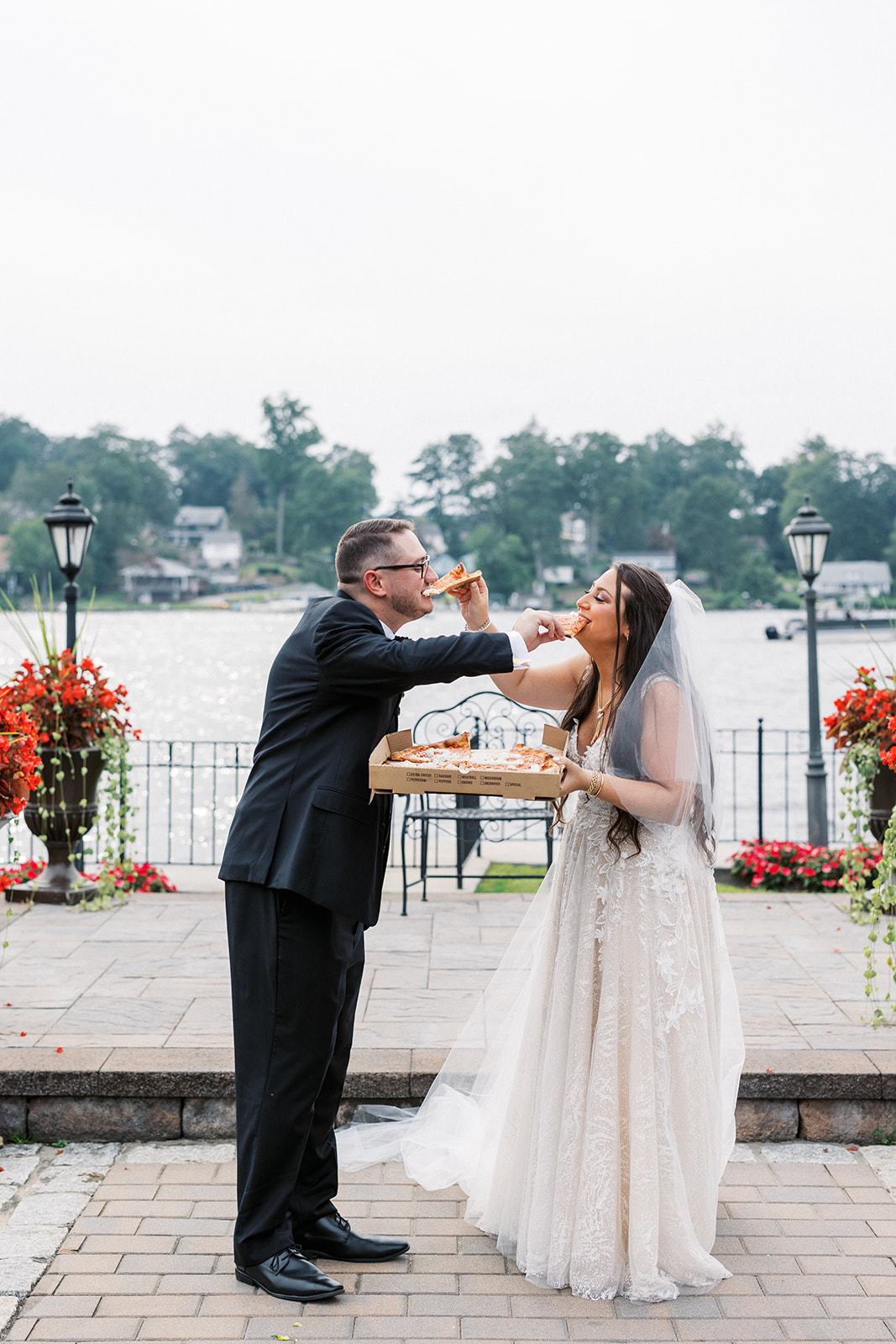 Newlyweds feed each other pizza while standing on a waterfront path at Villa Barone Hilltop Manor