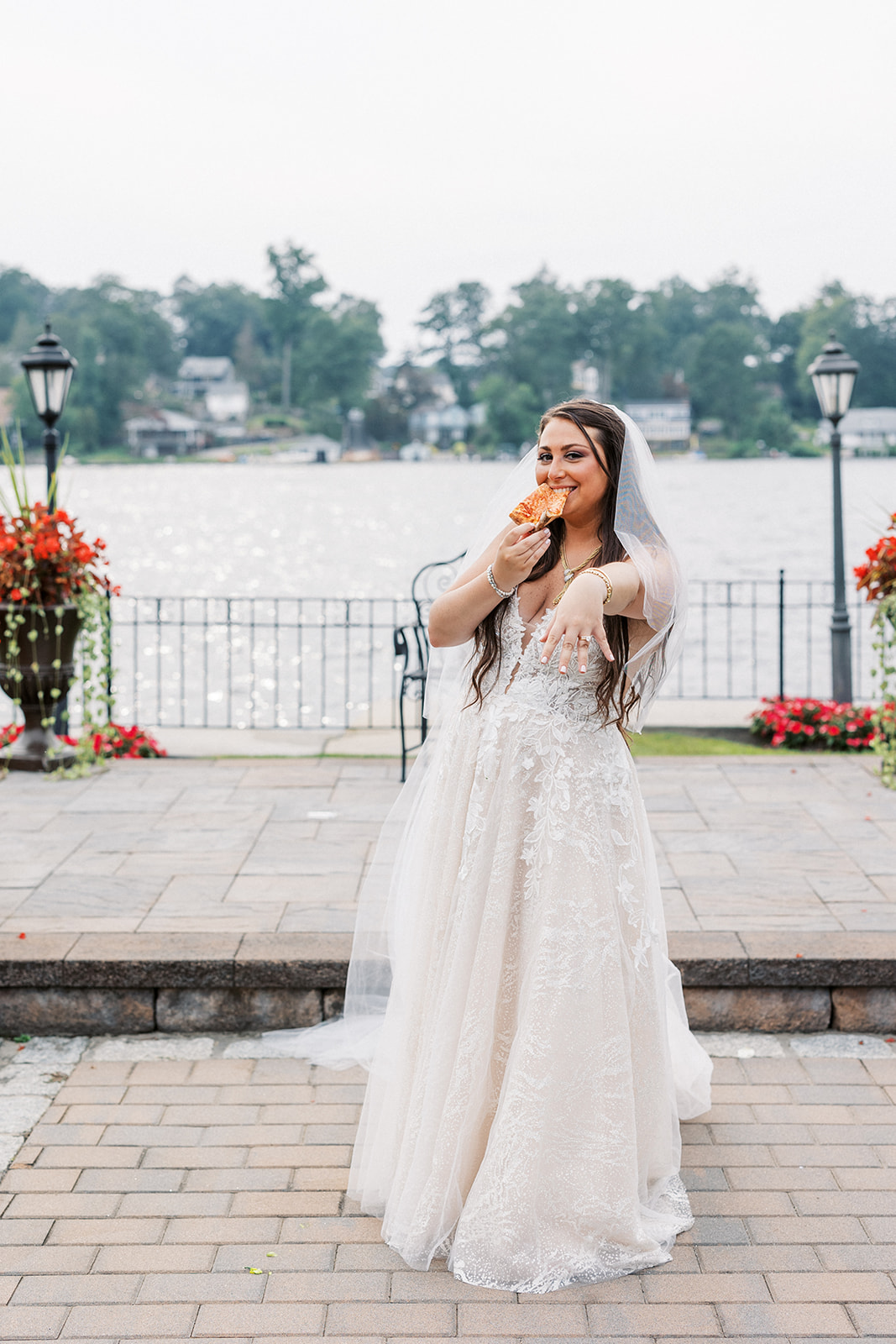 A bride displays her ring while eating a slice of pizza on a waterfront path at a Villa Barone Hilltop Manor