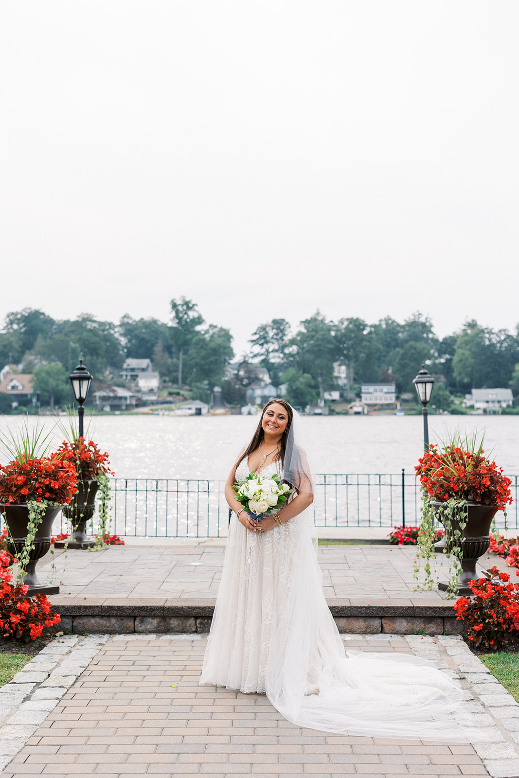 A bride holds her bouquet while standing with a long train on a waterfront brick path surrounded by red flowers