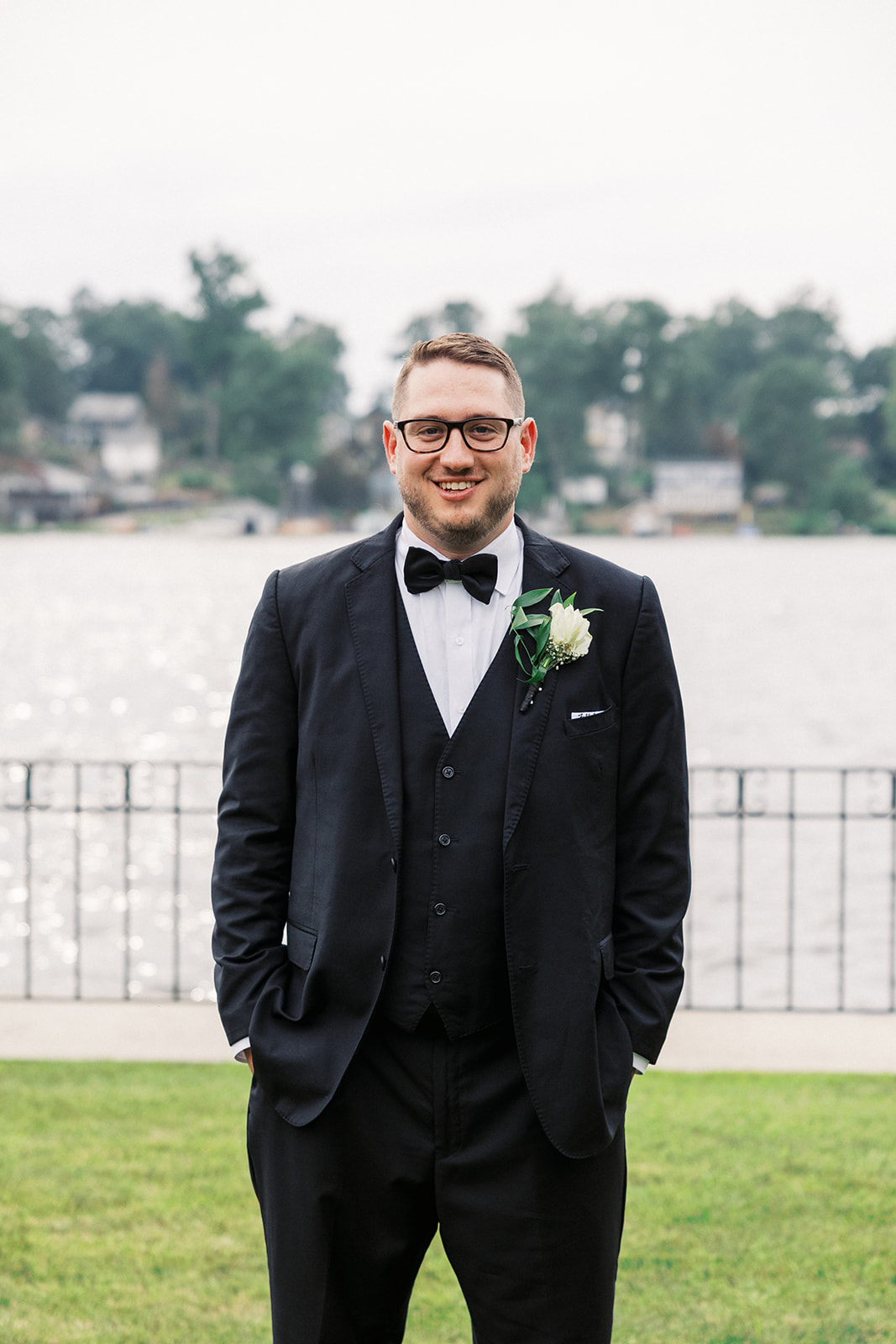 A groom in a black tuxedo stands with hands in pocket by the waterfront
