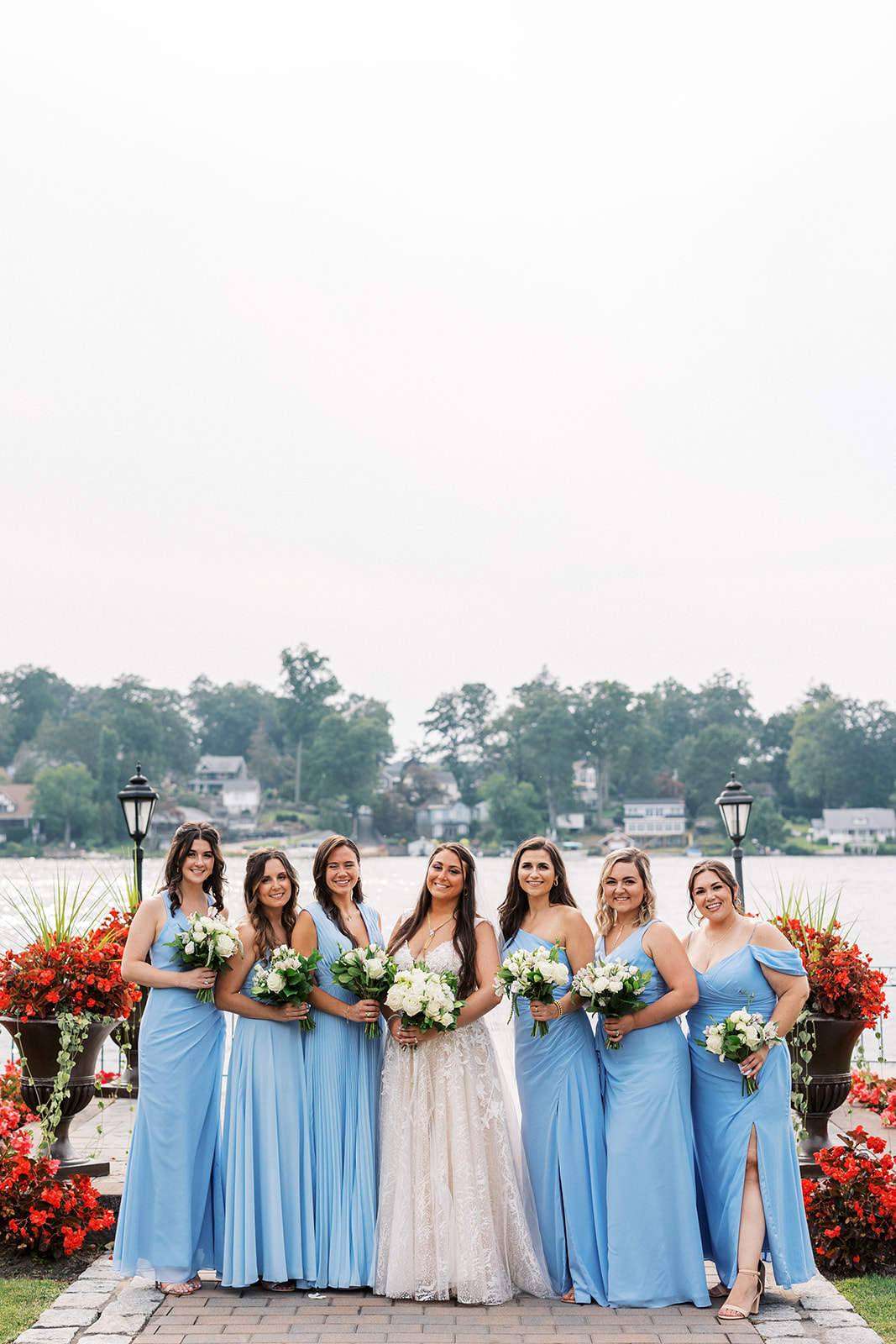 A bride stands with her bridal party in blue dresses surrounded by red flowers on the waterfront