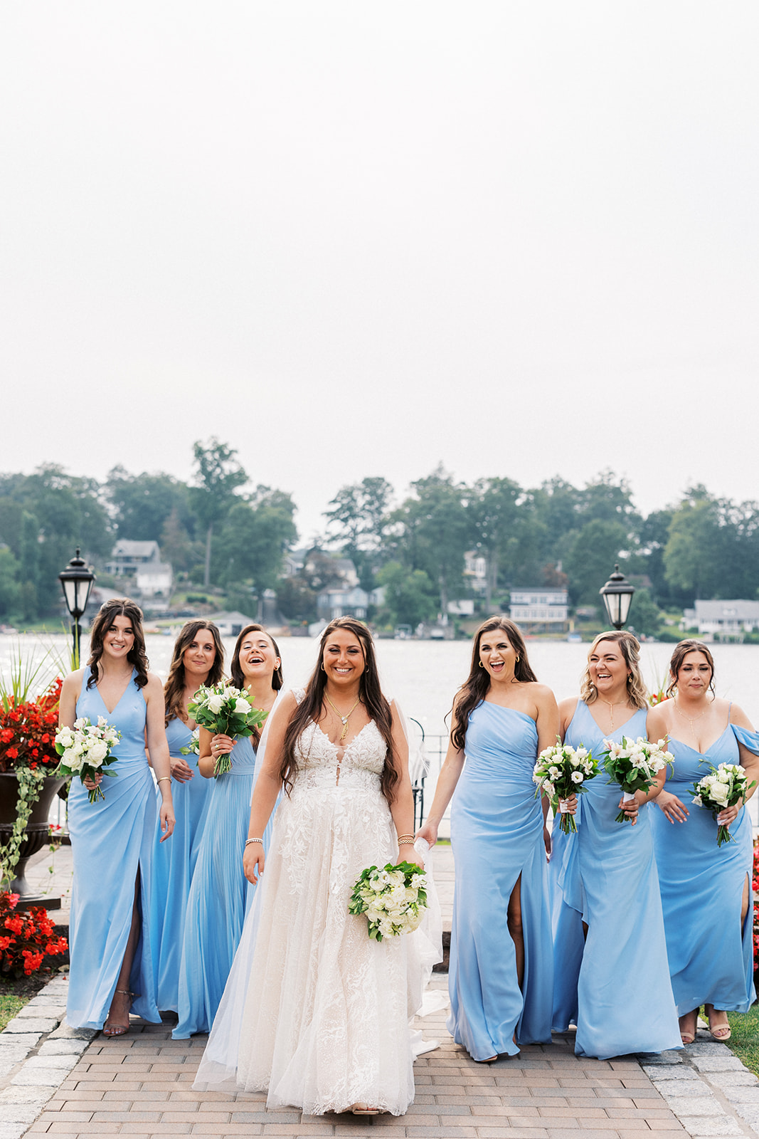 A bride walks up a brick paved path by the water with her bridal party in blue dresses behind her