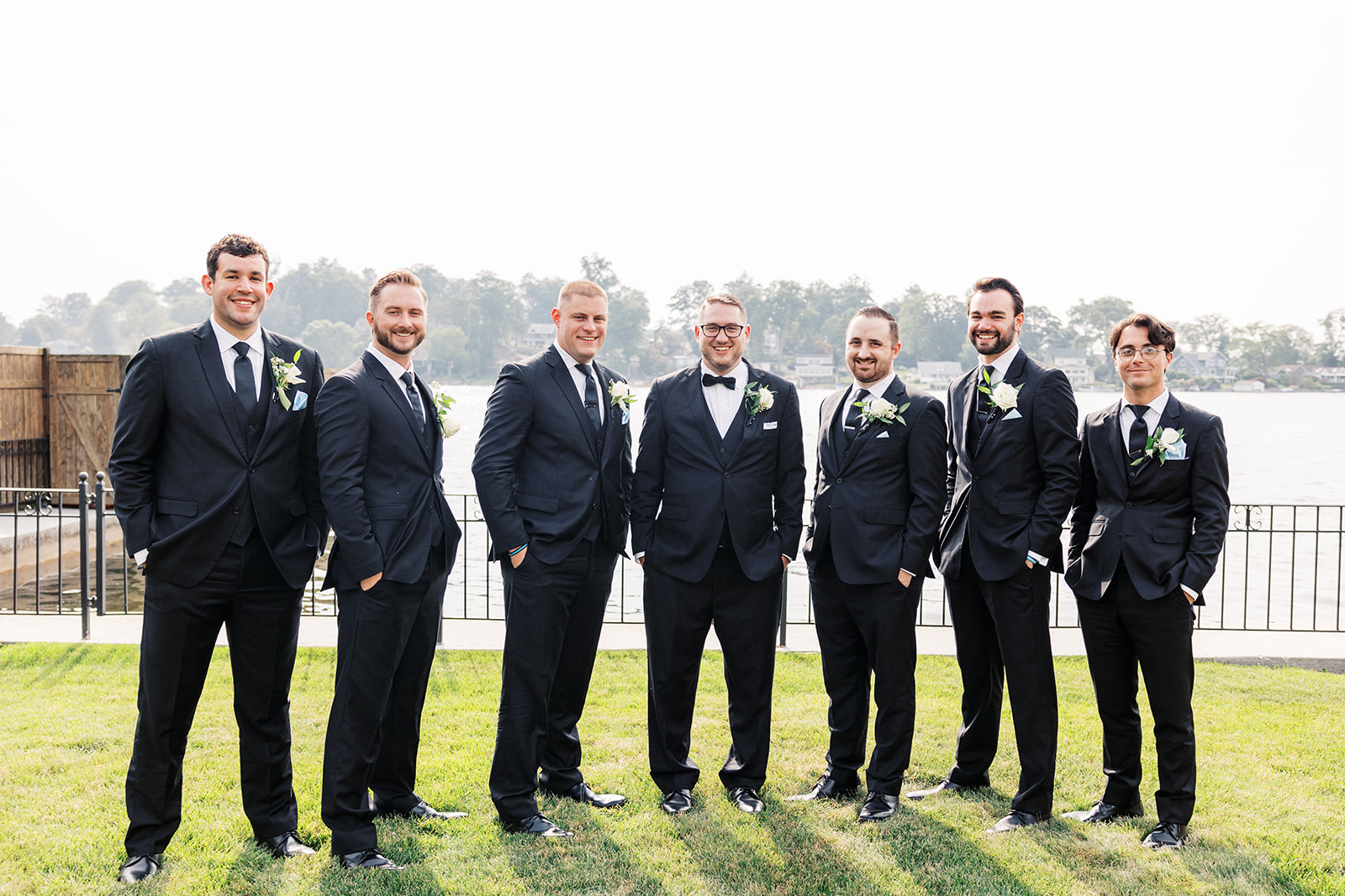 A groom stands with his wedding party on the waterfront in matching black suits