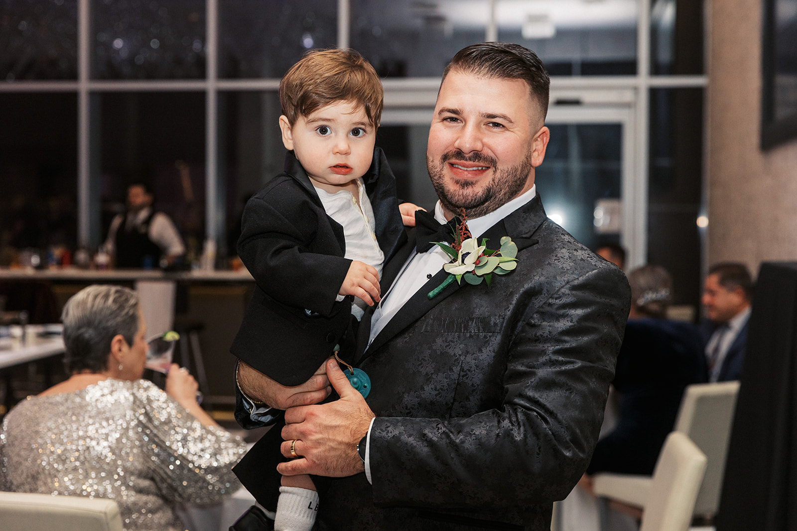 A groom in a black tuxedo holds a toddler boy during the reception of his Above Weddings