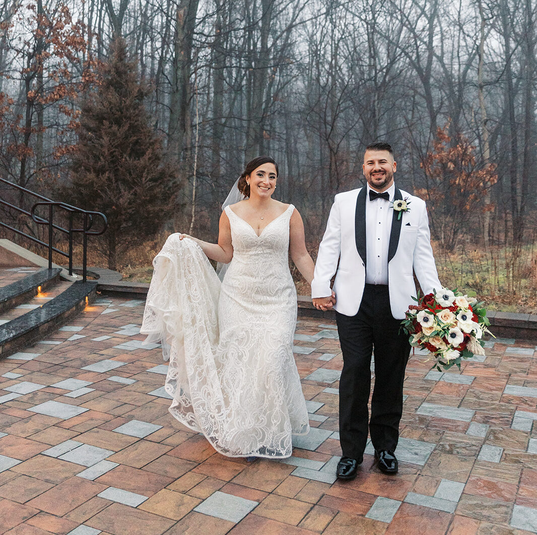 Newlyweds walk hand in hand on a brick patio in a white tuxedo and lace dress at their Above Weddings