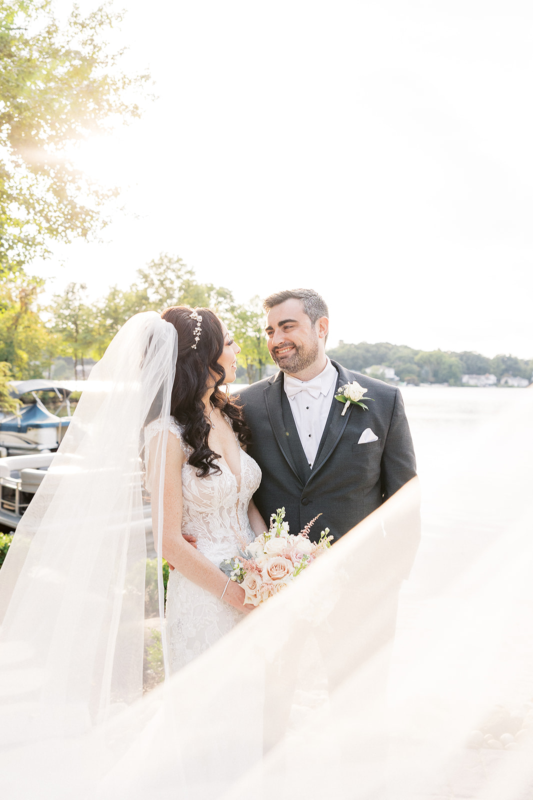 Newlyweds stand together smiling at each other on a waterfront patio while the veil blows around them in the wind