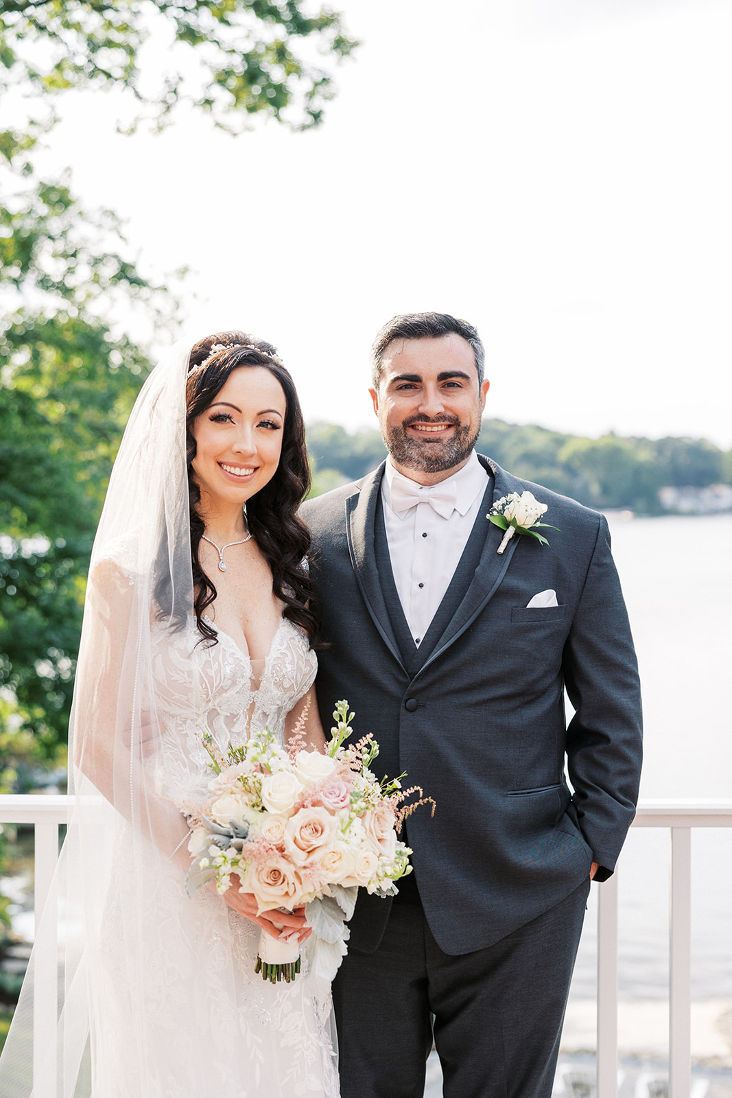 Newlyweds stand together by a lake on a patio holding a pink and white rose bouquet