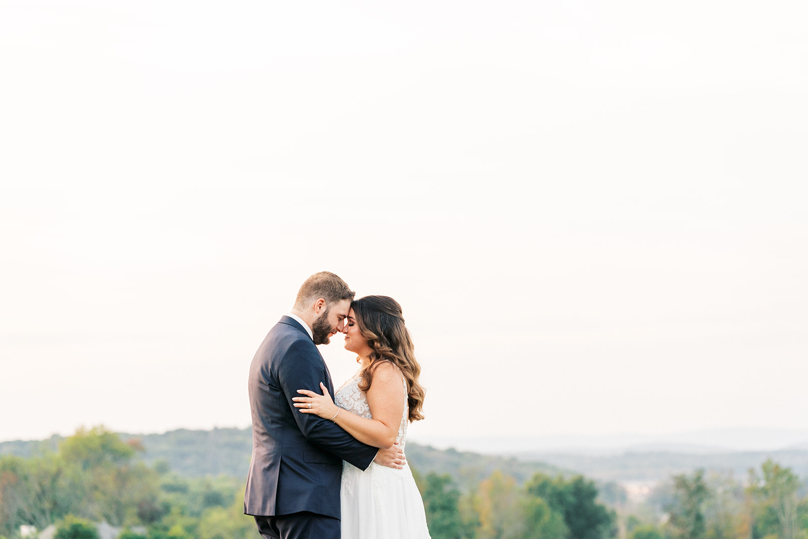Newlyweds touch foreheads while standing on a hill overlooking a valley at sunset