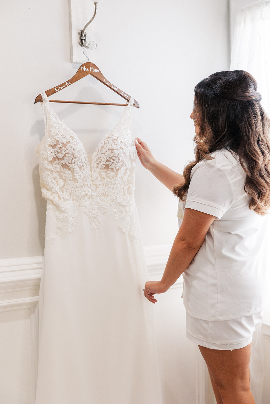A bride looks at her lace wedding dress handing on a wall by a window