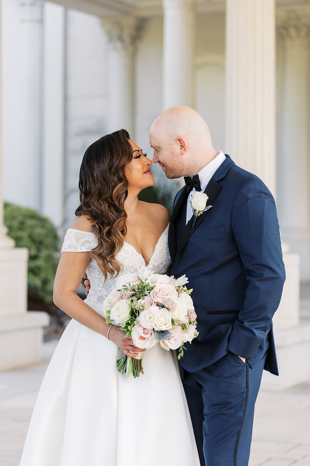 Newlyweds lean in for a kiss while standing in an ornate car port in a blue suit and white lace dress
