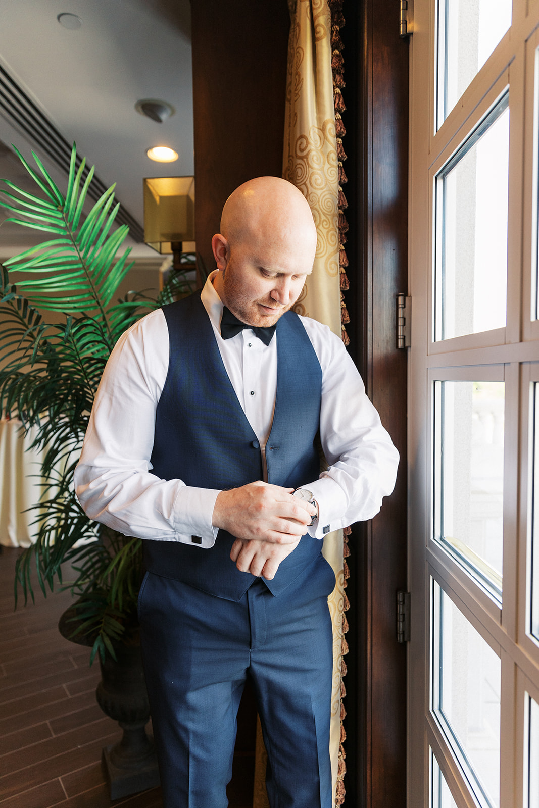 A groom in a blue suit checks his watch while standing in a window getting ready