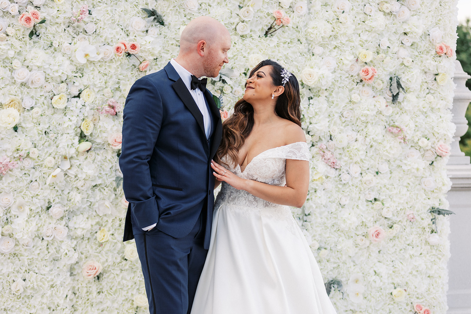 Newlyweds stand together smiling at each other in front of a wall of white and pink roses at their The Palace At Somerset Wedding