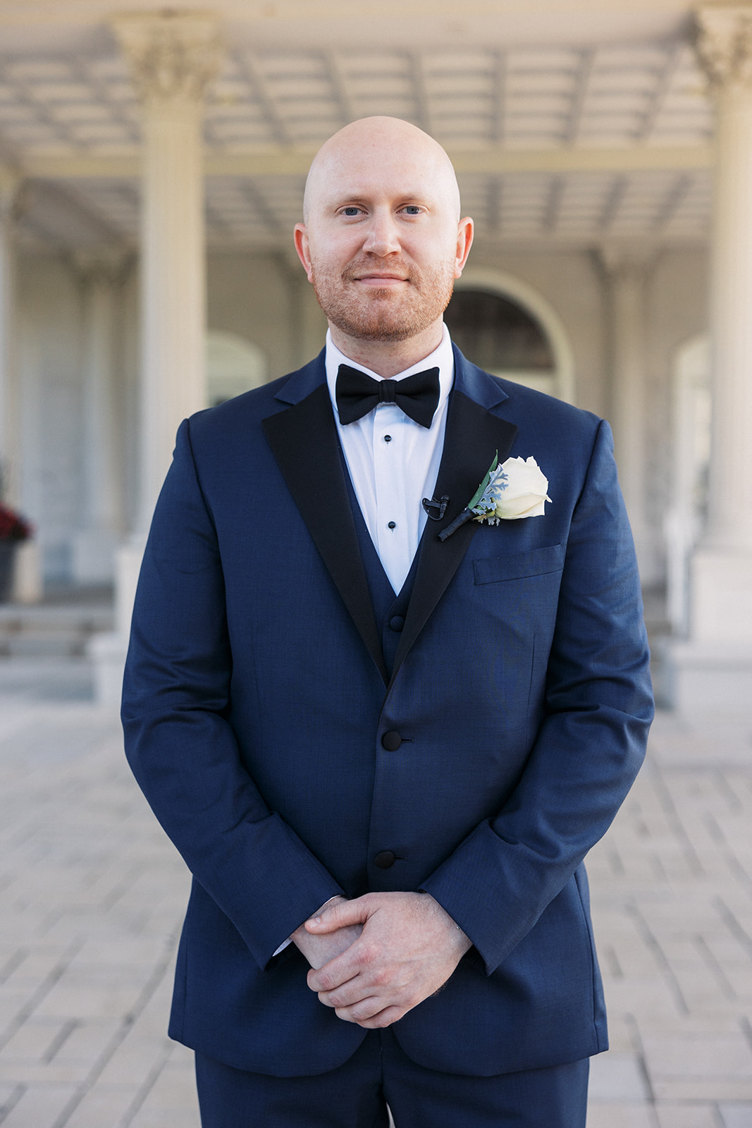 A groom in a blue suit stands with hands crossed in an ornate car port front entrance
