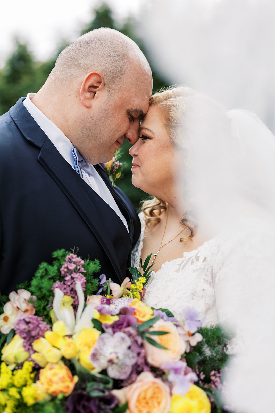 Newlyweds touch foreheads while standing in a garden holding a large colorful bouquet at their Forest Lodge Wedding