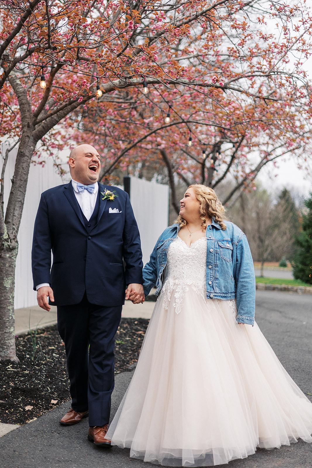 Newlyweds laugh while holding hands and walking down a sidewalk under blooming trees at their Forest Lodge Wedding