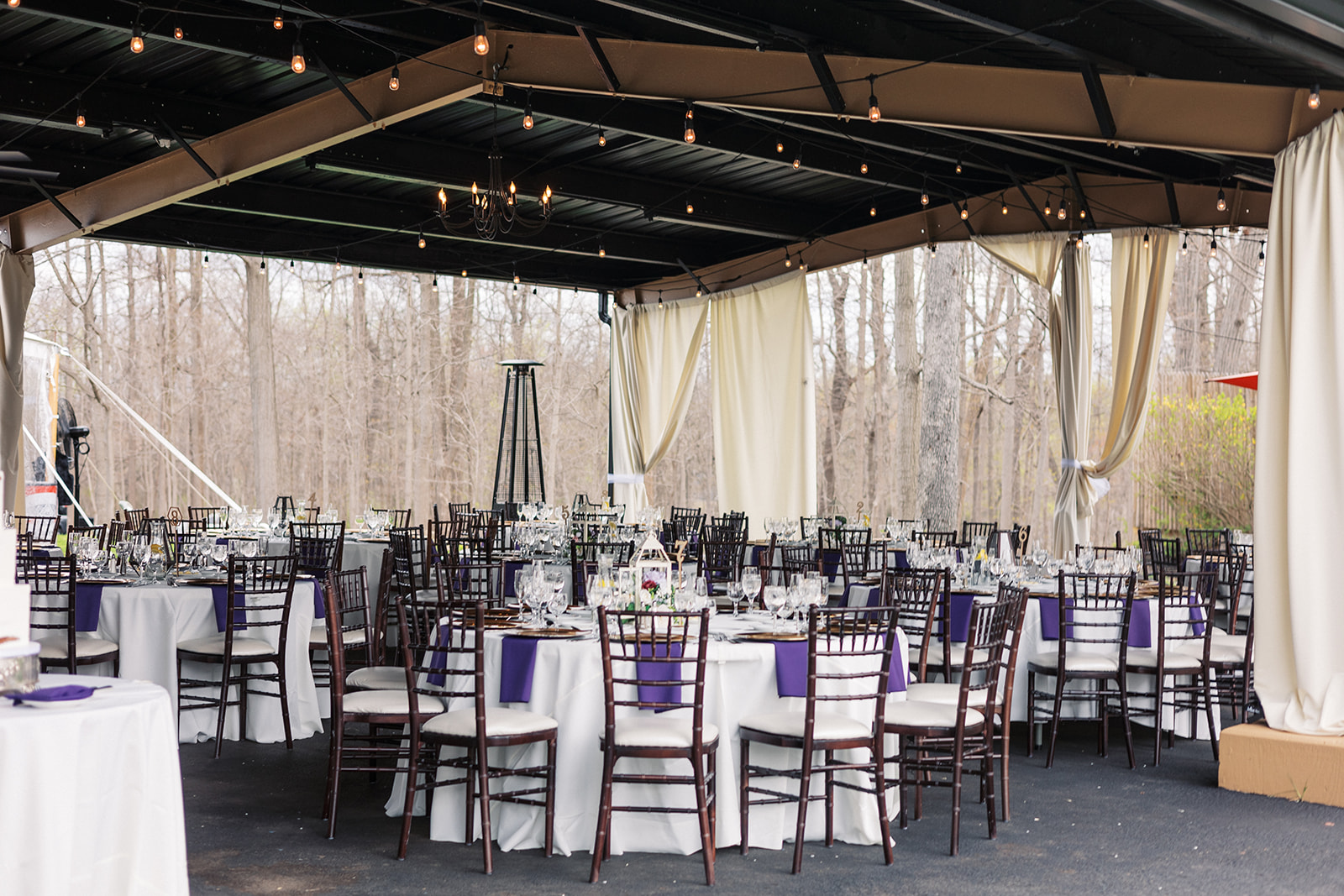 A Forest Lodge Wedding reception set up under an outdoor overhang with market lights and purple napkins