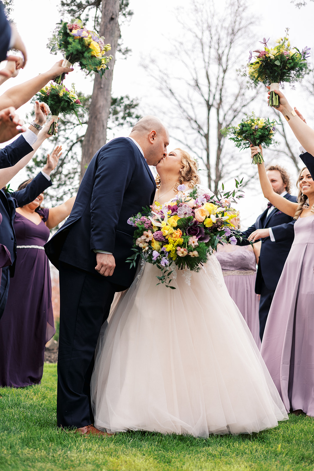 Newlyweds kiss while surrounded by celebrating wedding parties with bouquets raised