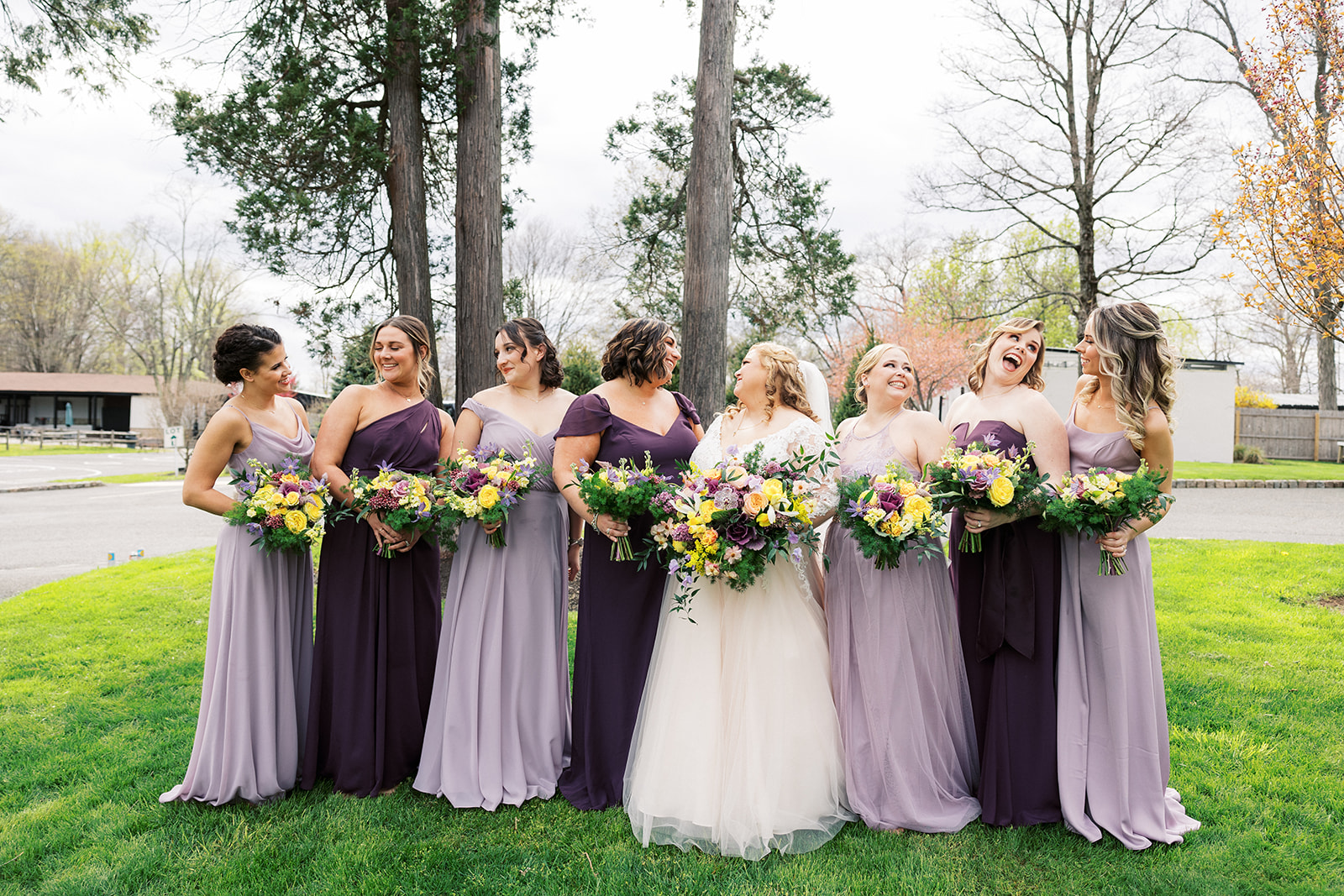 A bride stands in a grassy lawn under some trees laughing with her bridesmaids in purple dresses at her Forest Lodge Wedding