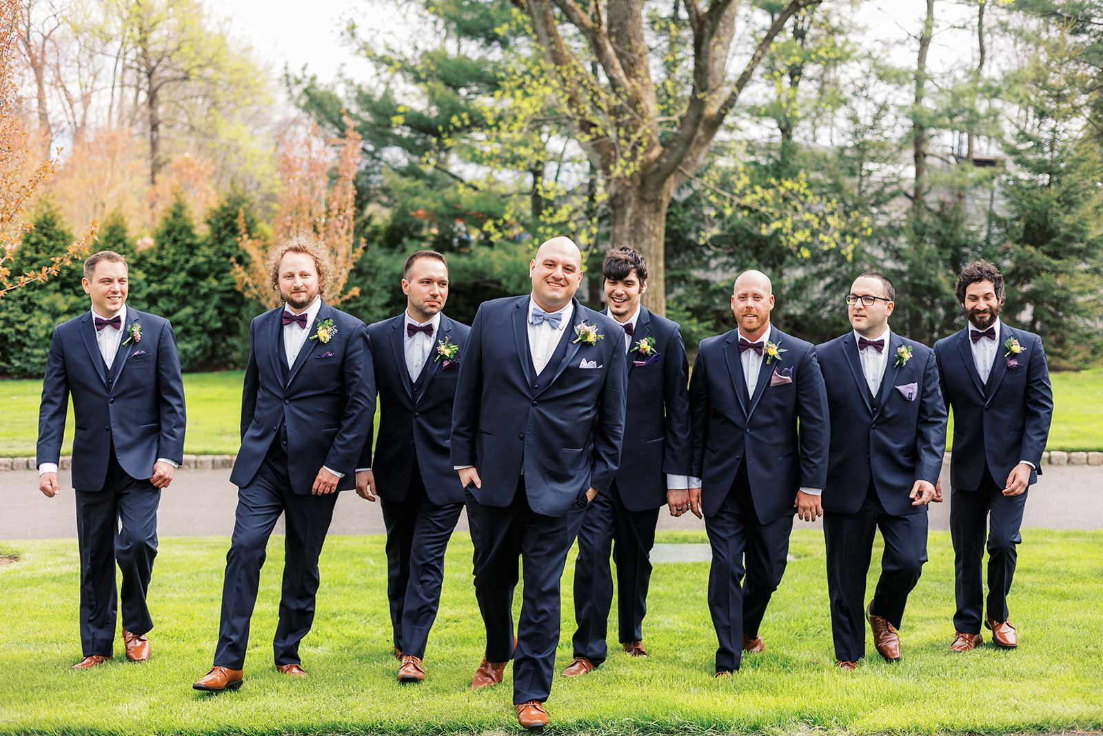 A groom in a blue suit walks with his matching groomsmen through a grassy lawn