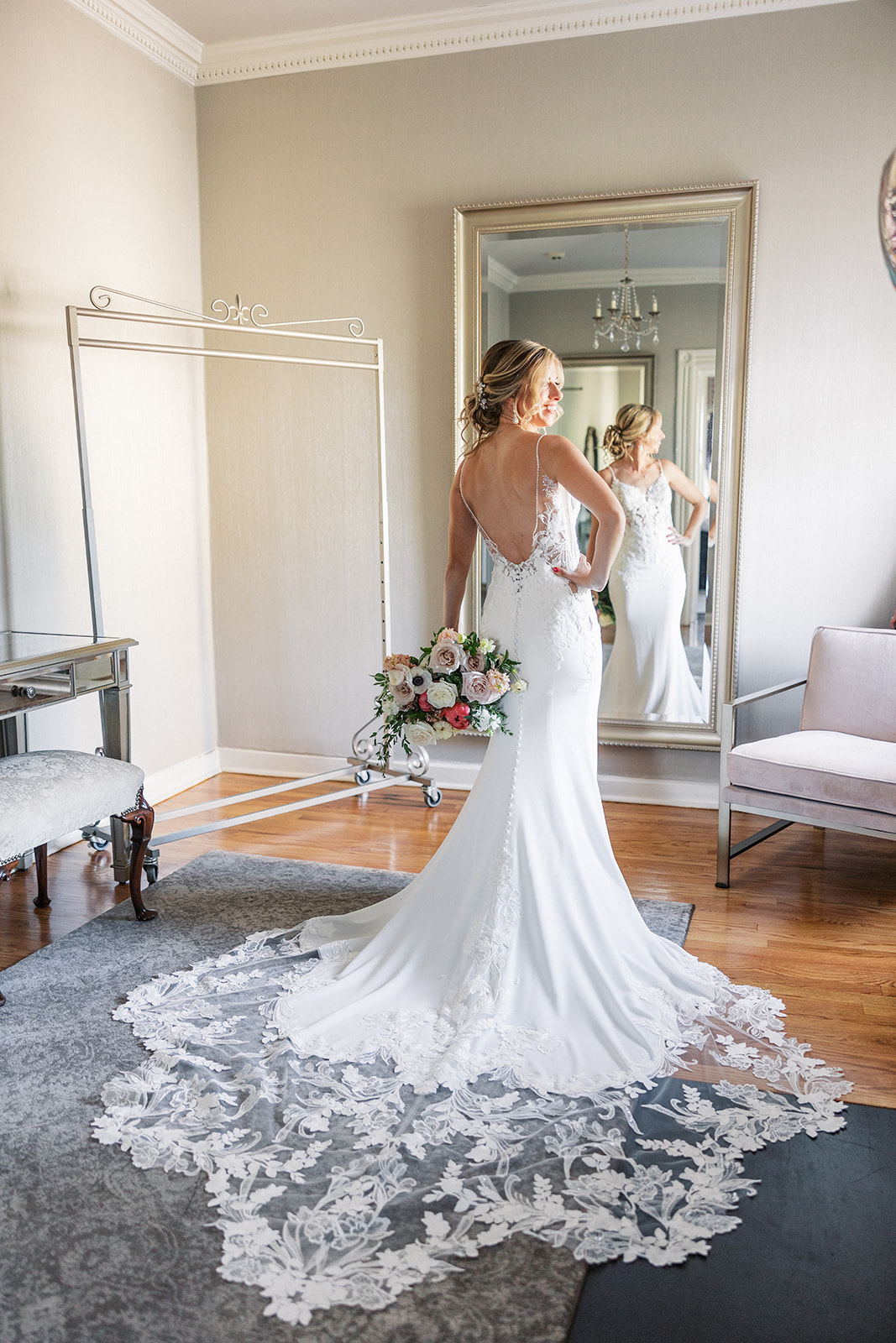 A bride stands with a hand on her hip in a mirror with her long lace train flowing behind her