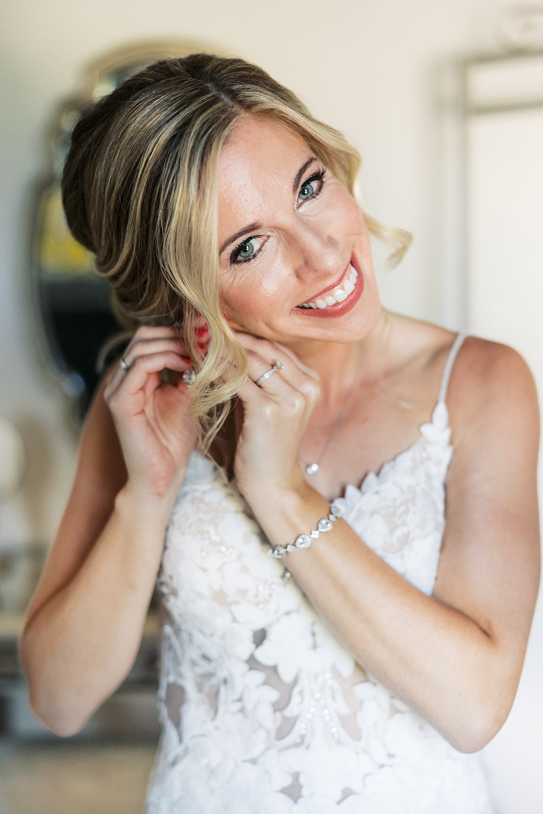 A bride smiles while putting on her earrings in a lace dress