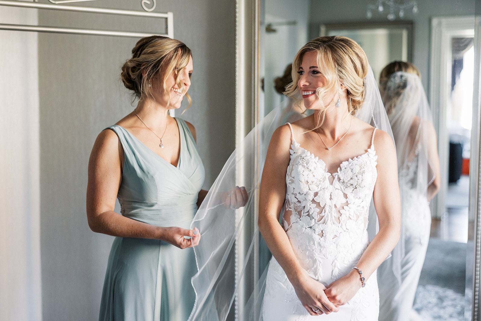A bride stands in a mirror getting ready with her bridesmaid