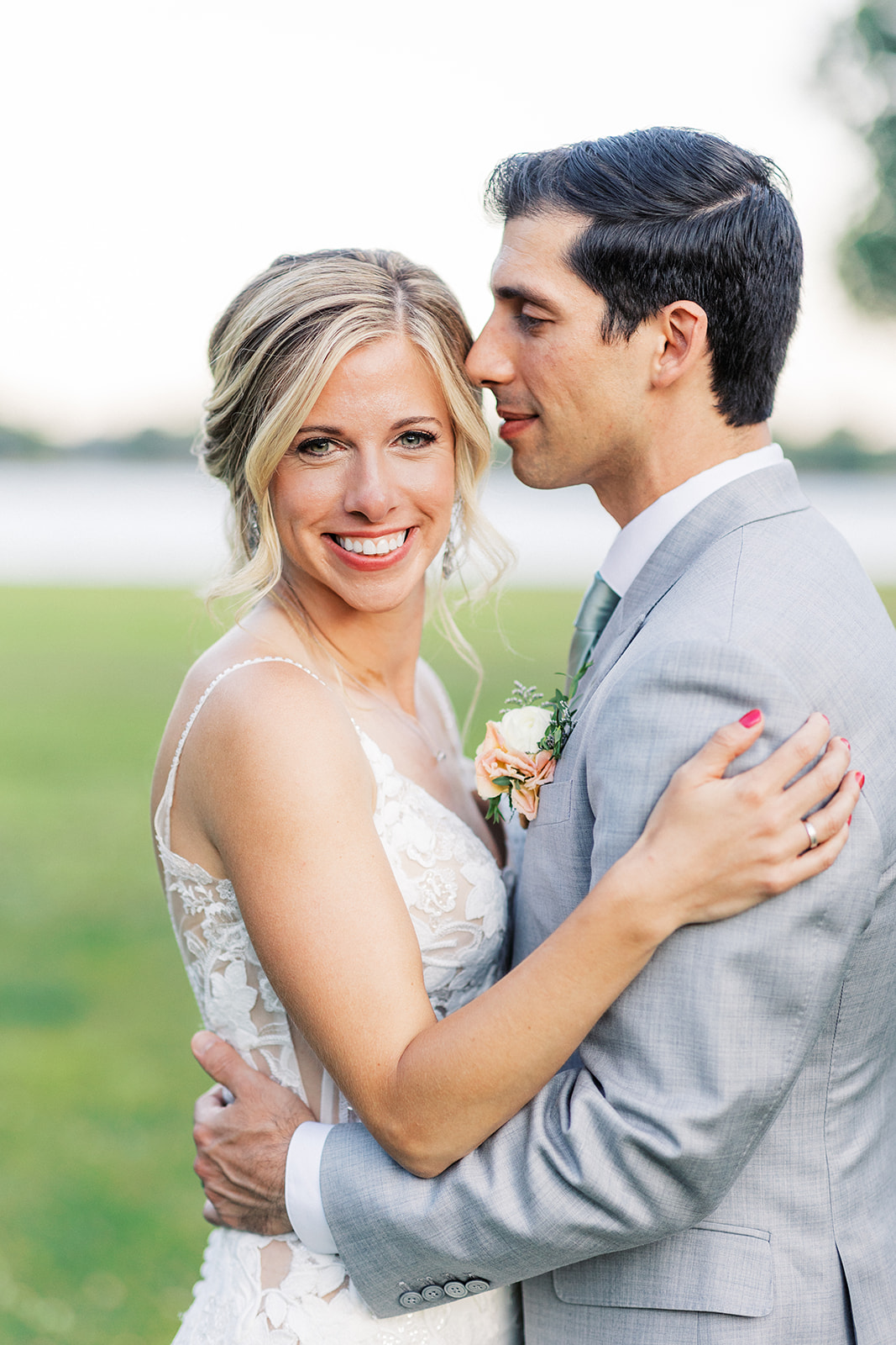 A bride smiles over her shoulder while standing in a lawn as her groom in a grey suit nuzzles her hair