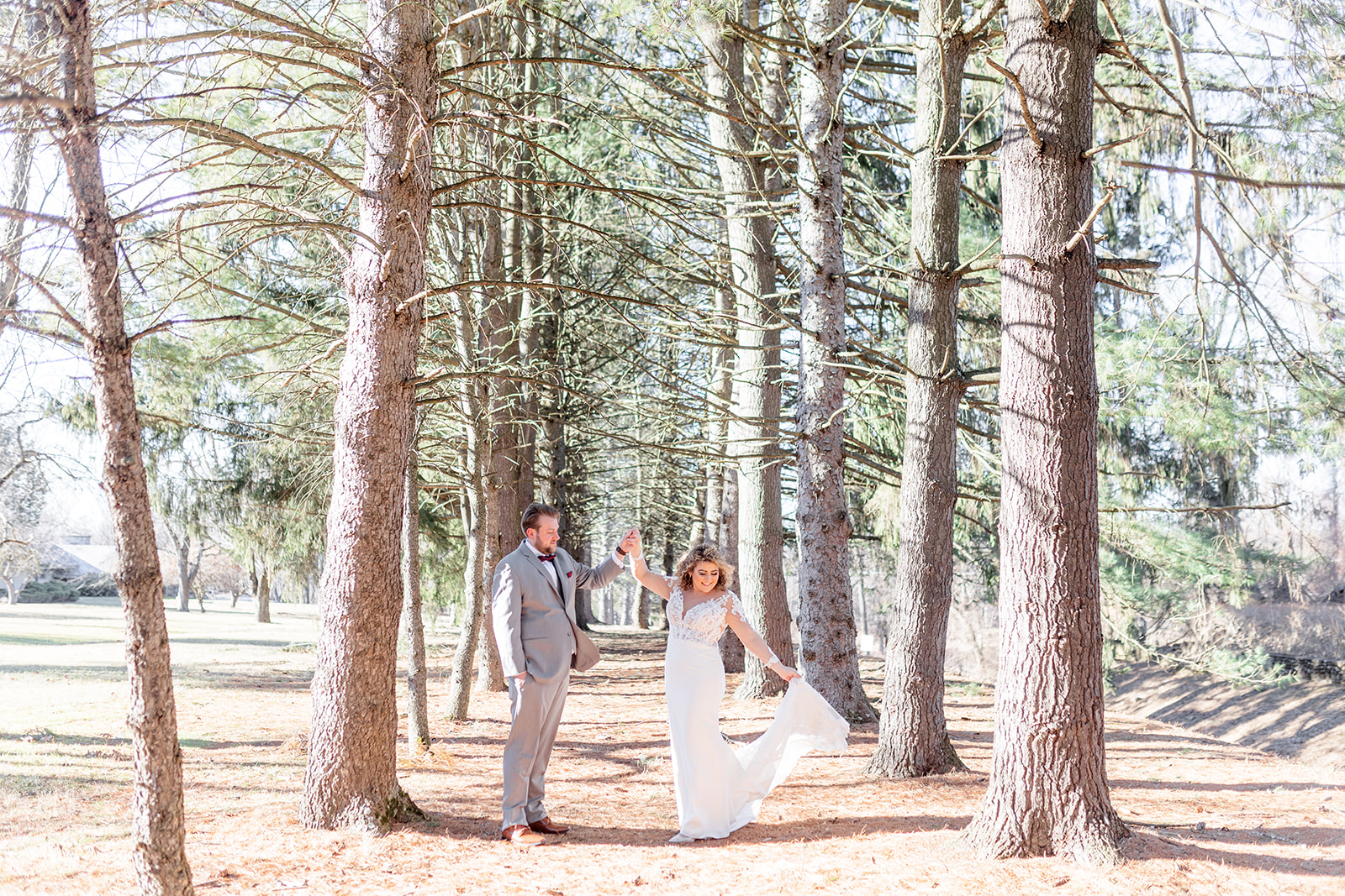 Newlyweds dance and twirl the dress in a line of tall pine trees at sunset at The Refinery At Perona Farms