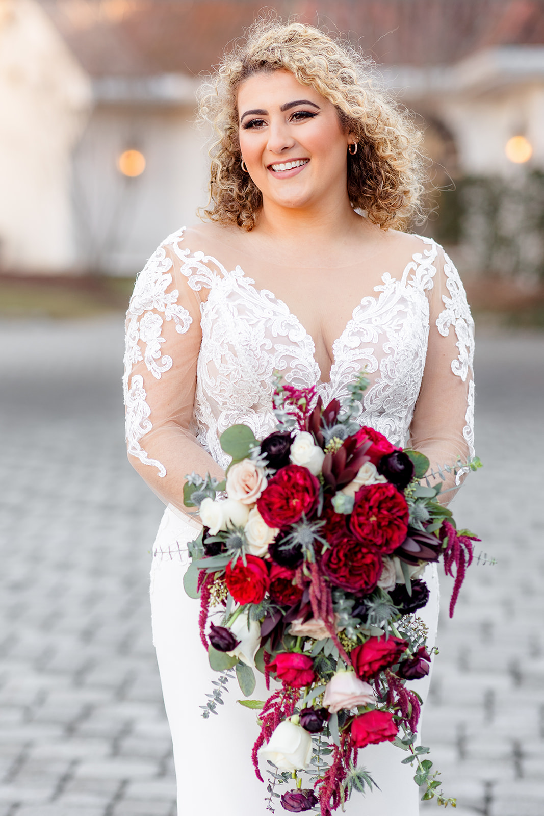 A bride smiles while standing in her lace dress holding her red bouquet on a paved driveway at sunset