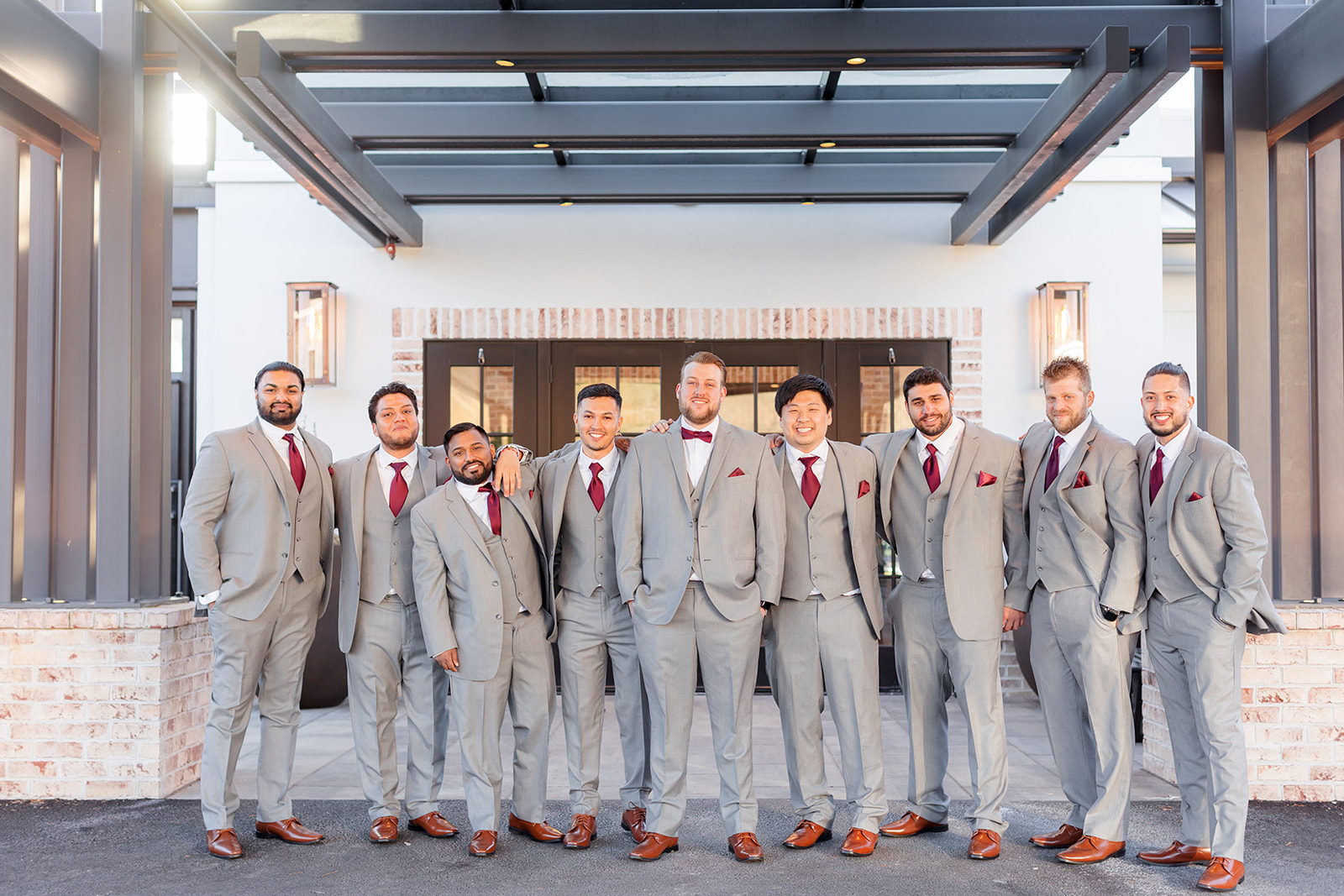 A groom stands with his many groomsmen in grey suits and red ties at the entrance to the wedding venue