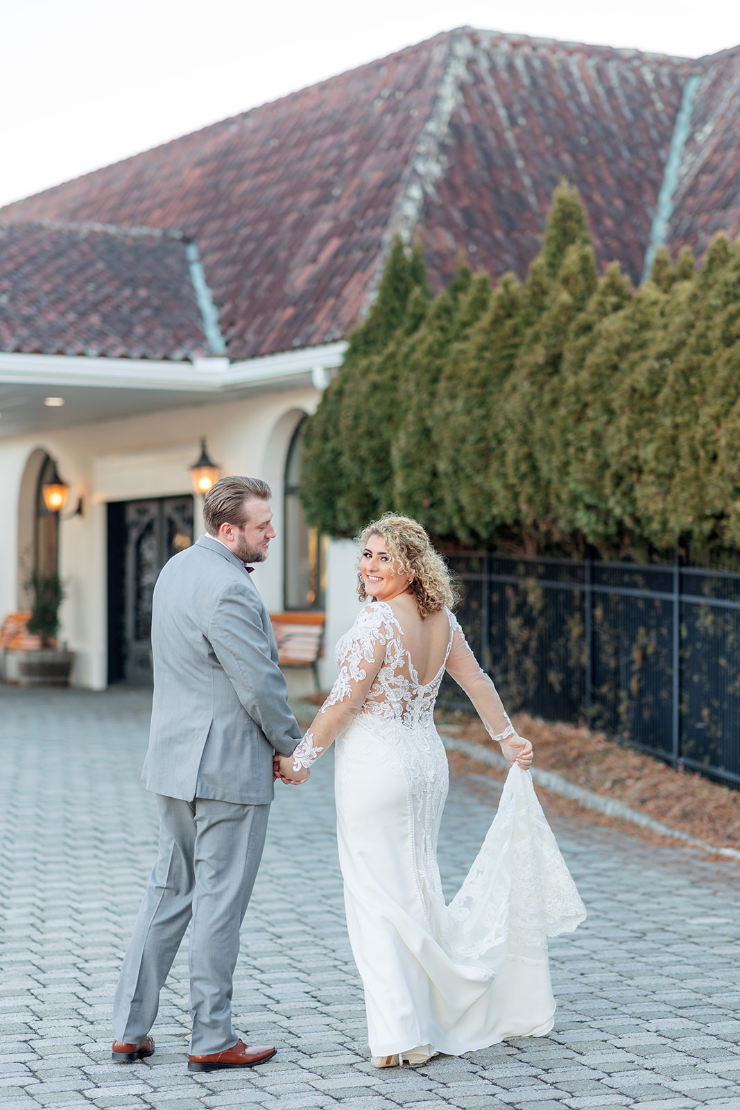Newlyweds hold hands while walking on a grey brick paved driveway to their wedding reception